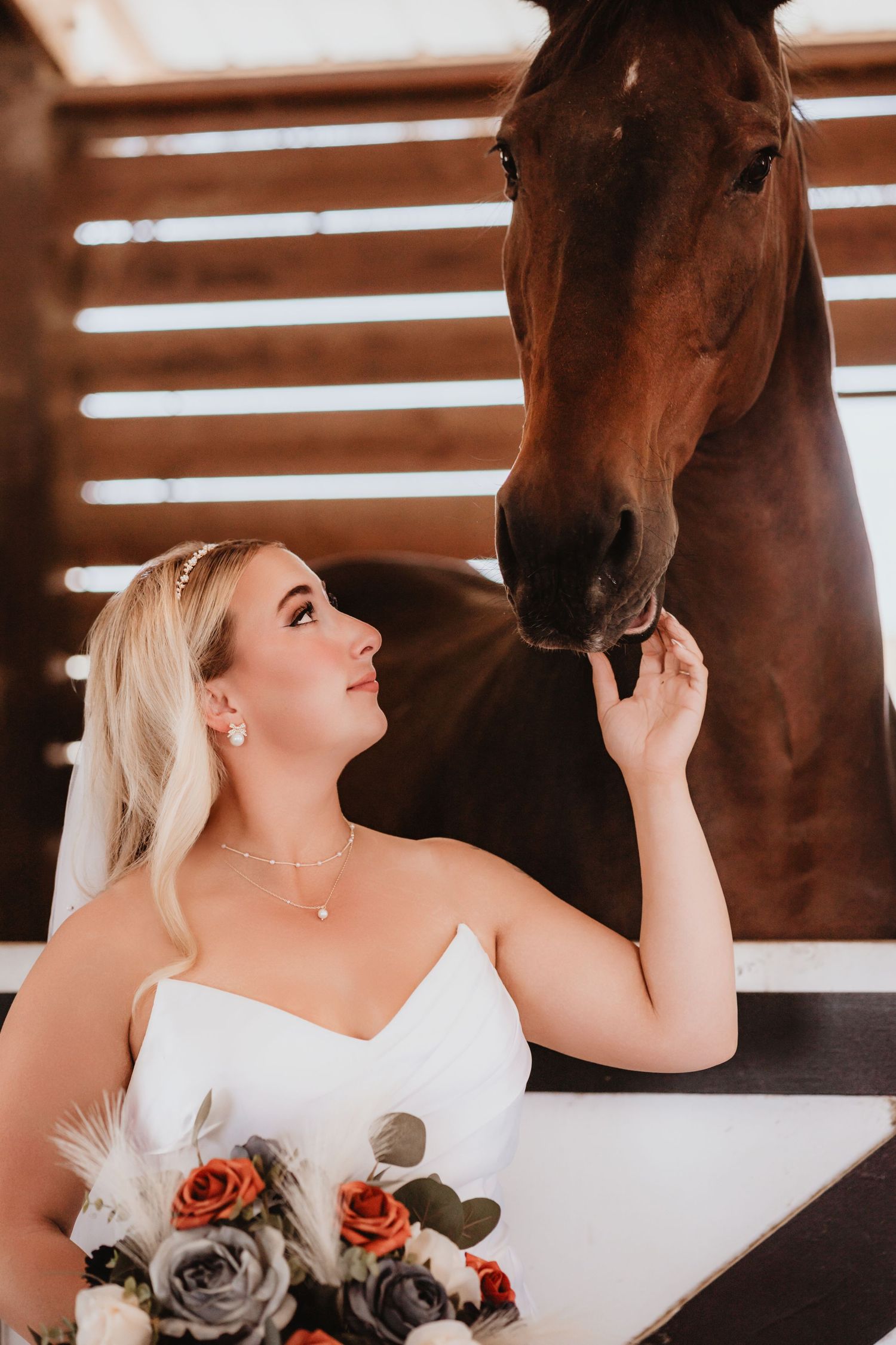 Bride in white dress gazes up at a brown horse while holding a colorful wedding bouquet.
