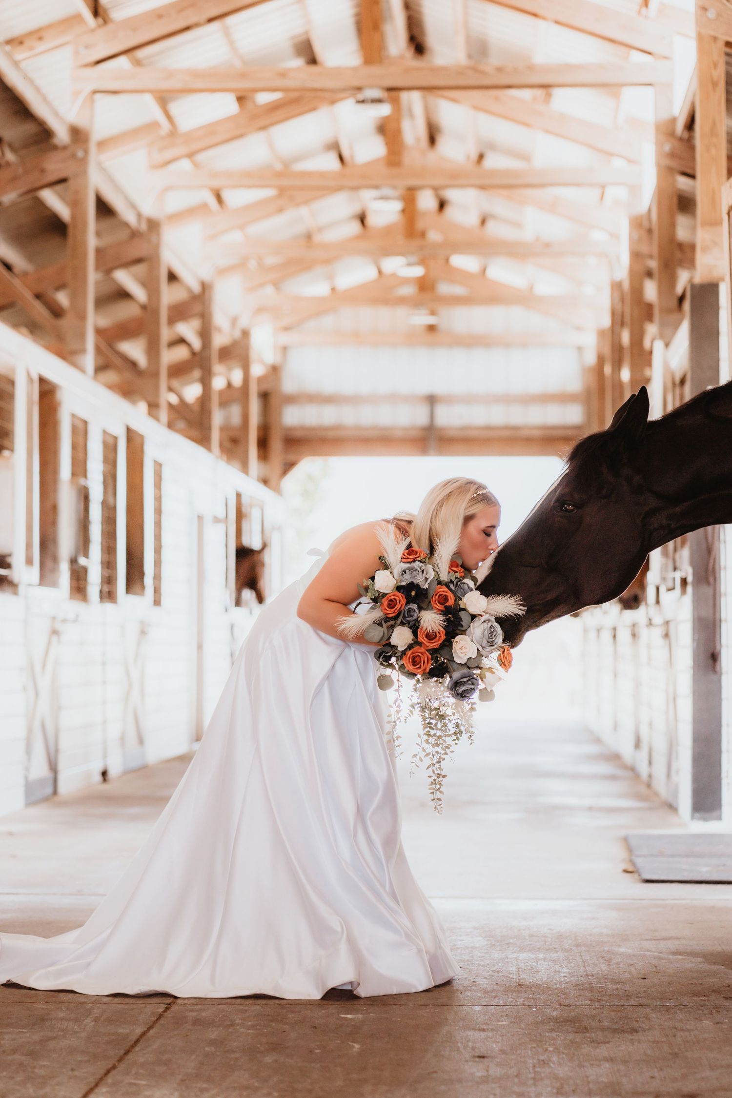 Blonde bride in white gown shares a tender moment with a black horse in a barn.