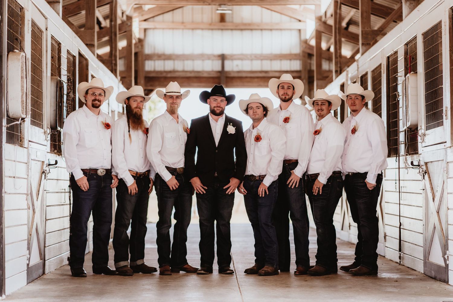 Groom in black suit and cowboy hat poses with groomsmen in white shirts and cowboy hats inside a barn.