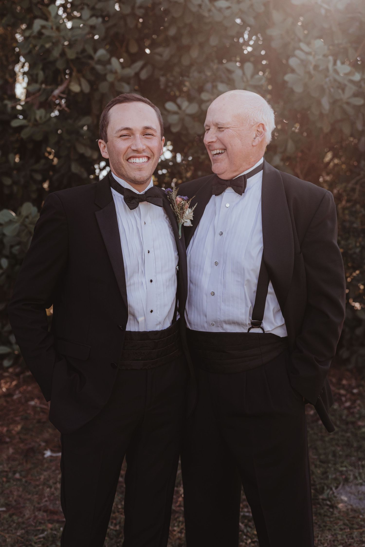 Two men in black tuxedos with bow ties laugh together outdoors at a wedding celebration.