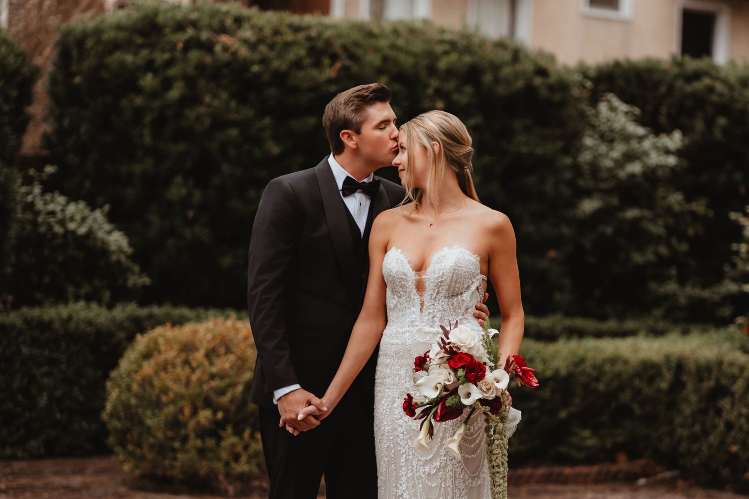 Groom kisses brides cheek as she smiles holding a red and white bouquet in front of green hedges.