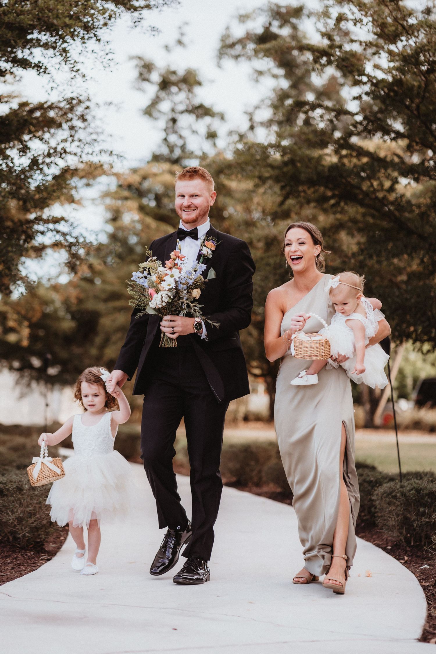 Bridesmaid in sage gown walks with groomsman and two young flower girls down the aisle.