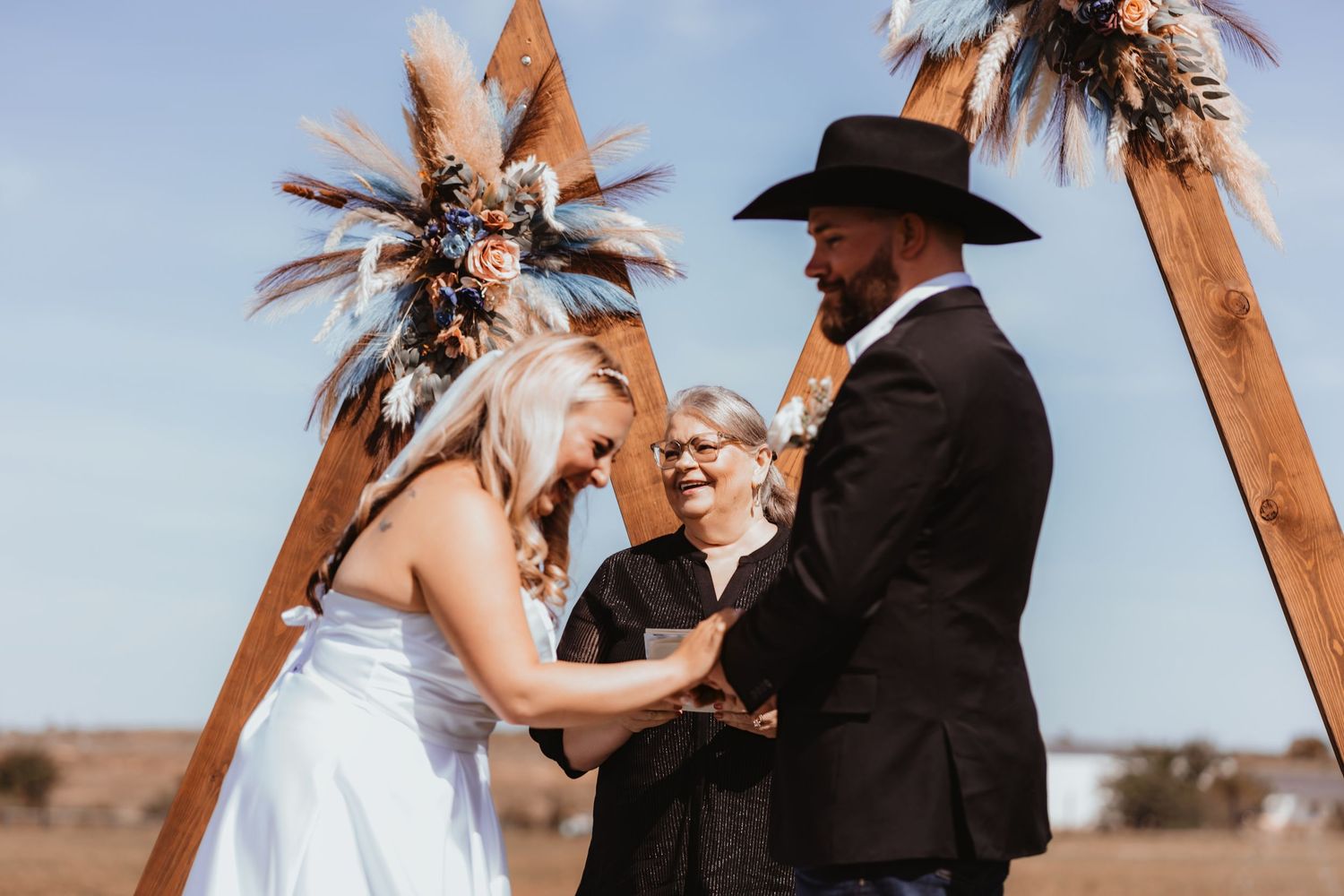 Bride and groom laugh joyfully during outdoor wedding ceremony at rustic boho wooden triangular arch altar.