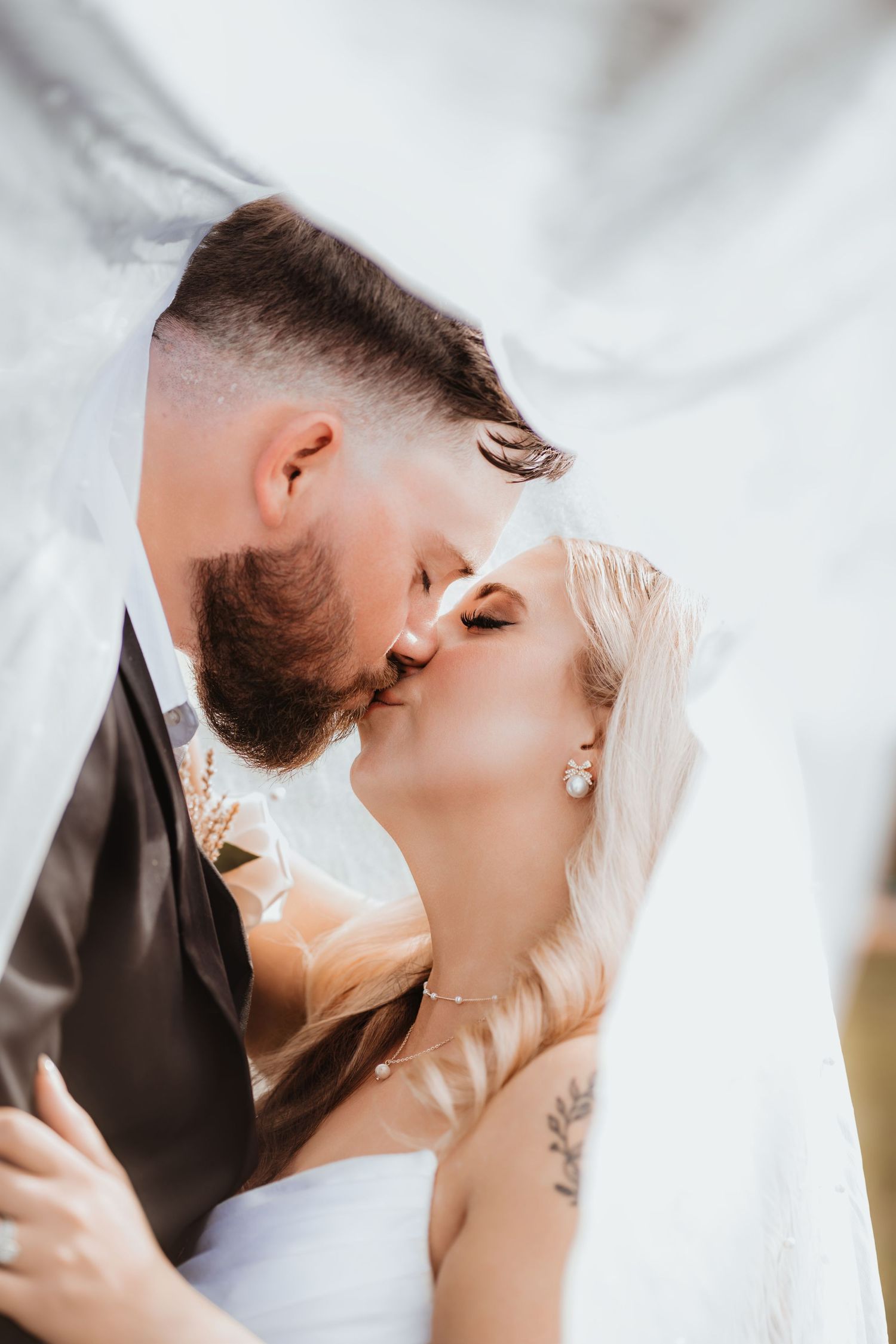 Groom kisses bride passionately under a flowing white veil, bride adorned with diamond earrings and floral tattoo.