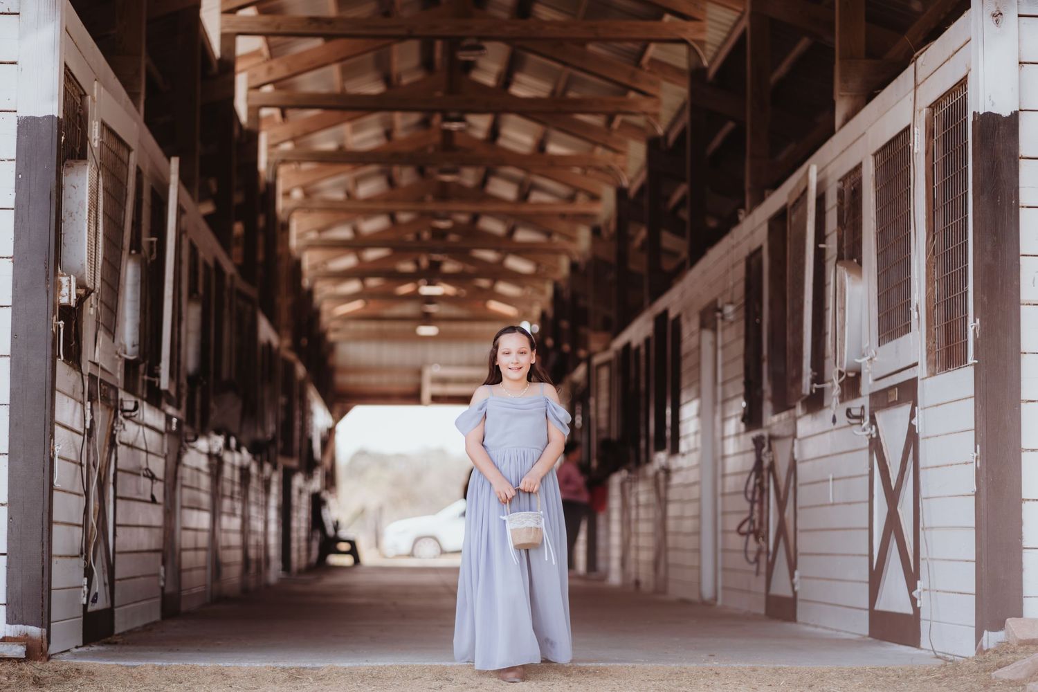 Young flower girl in a light blue gown posing in a long wooden horse stable corridor at a wedding.