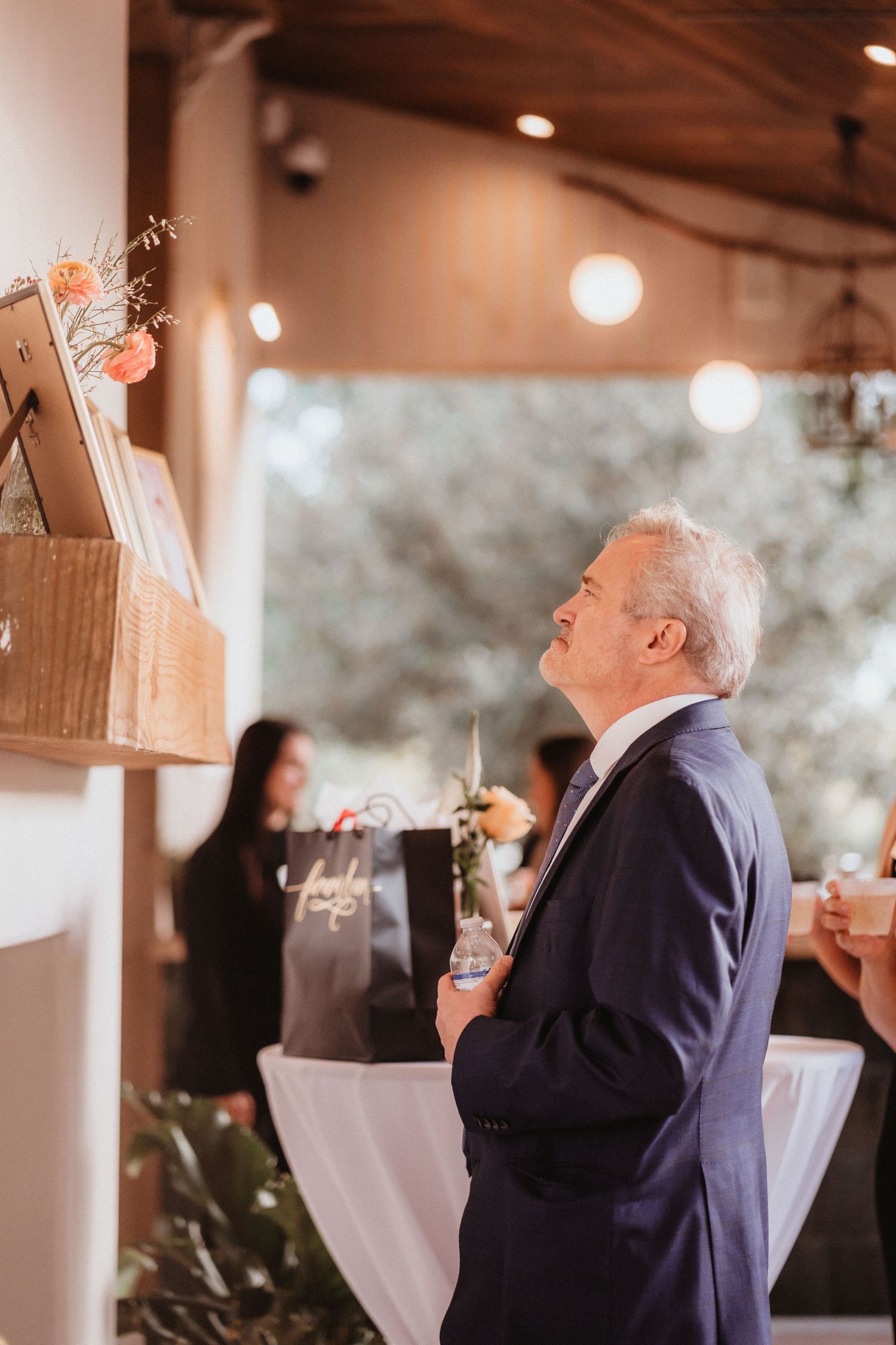 Elderly man in navy suit gazing emotionally at framed wedding photos displayed on rustic wooden shelf.