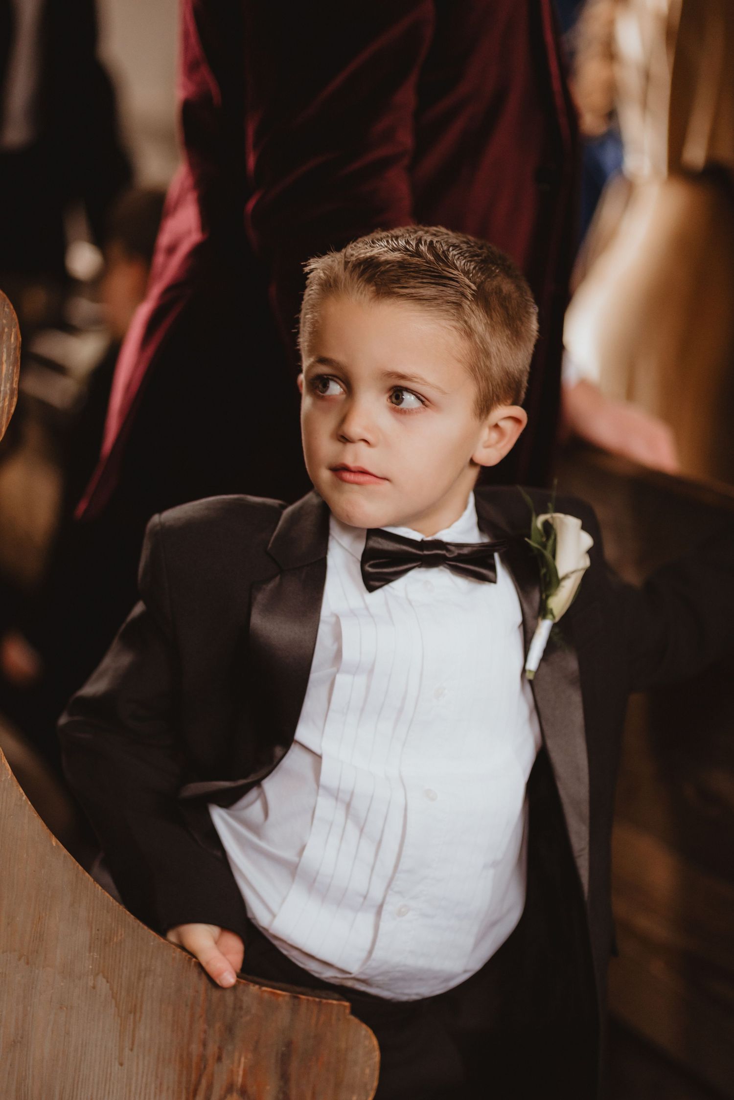 Adorable young ring bearer in black tuxedo with bow tie and white boutonniere looks up attentively during wedding ceremony.