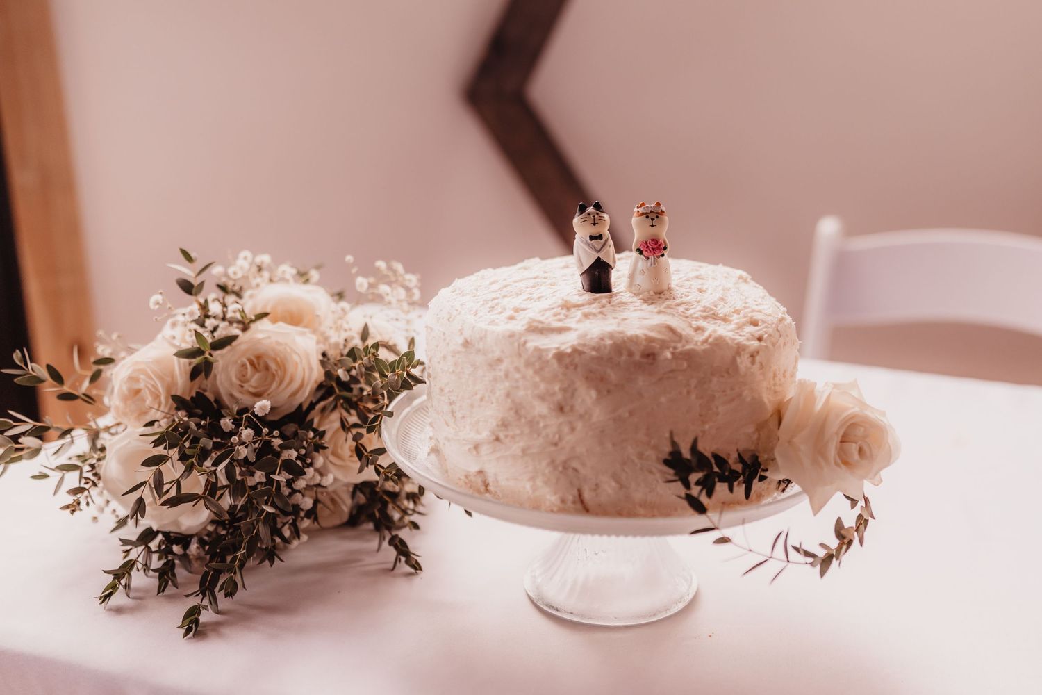 White wedding cake with bride and groom figurines beside a white rose bouquet on a stand.
