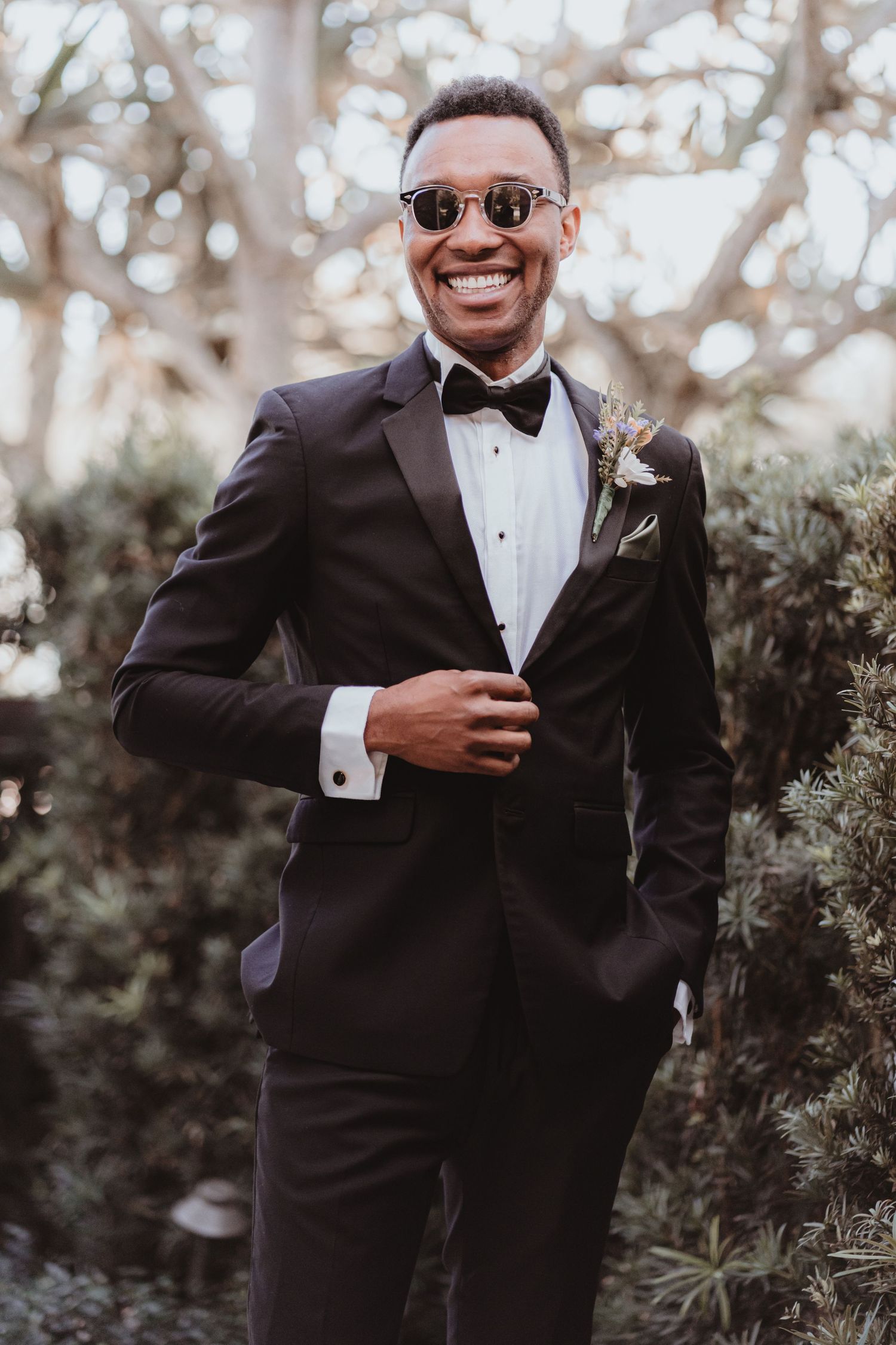 Groom in black tuxedo and sunglasses smiling confidently outdoors at his wedding.