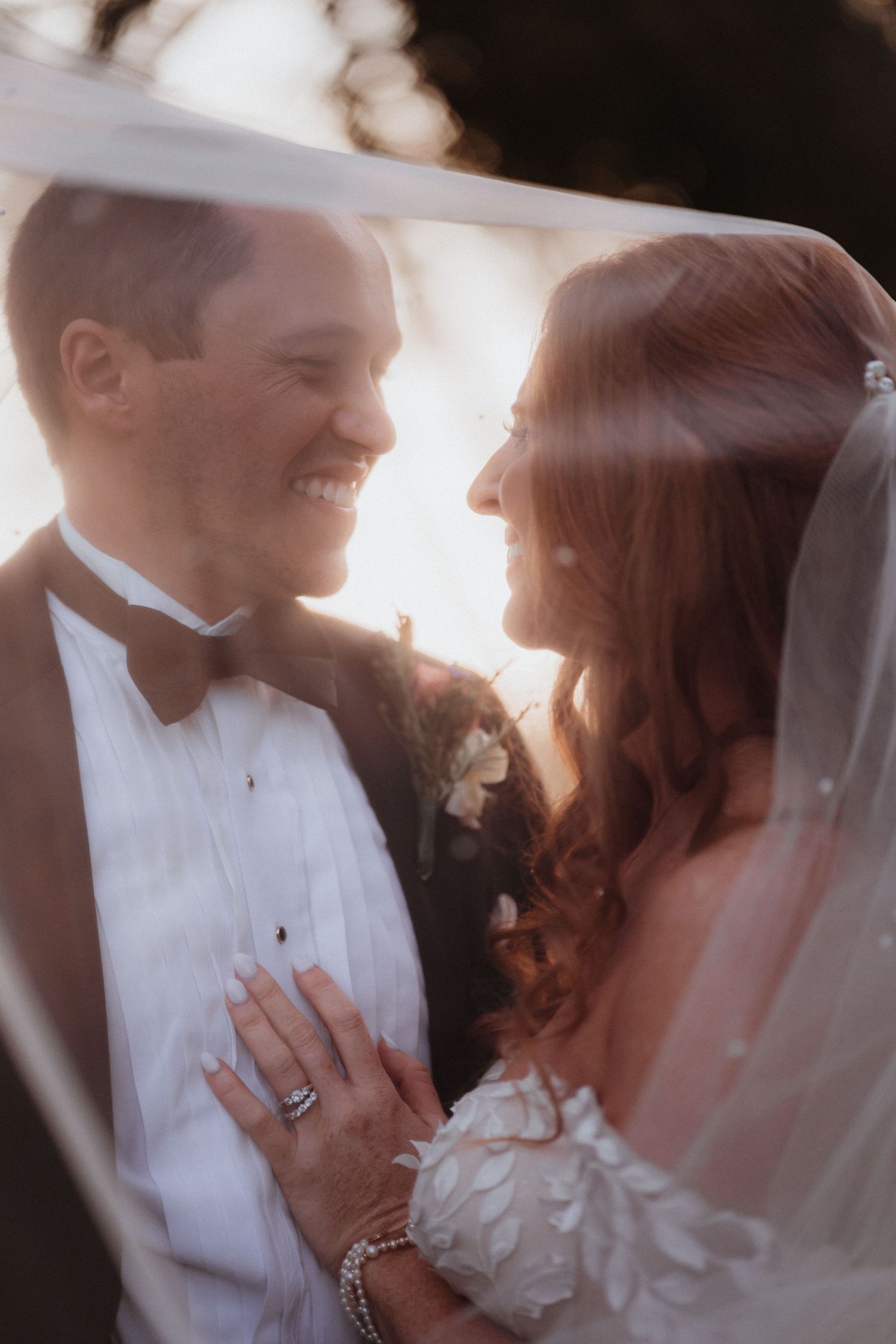 Smiling bride and groom gaze at each other under a flowing veil in warm golden sunset light.