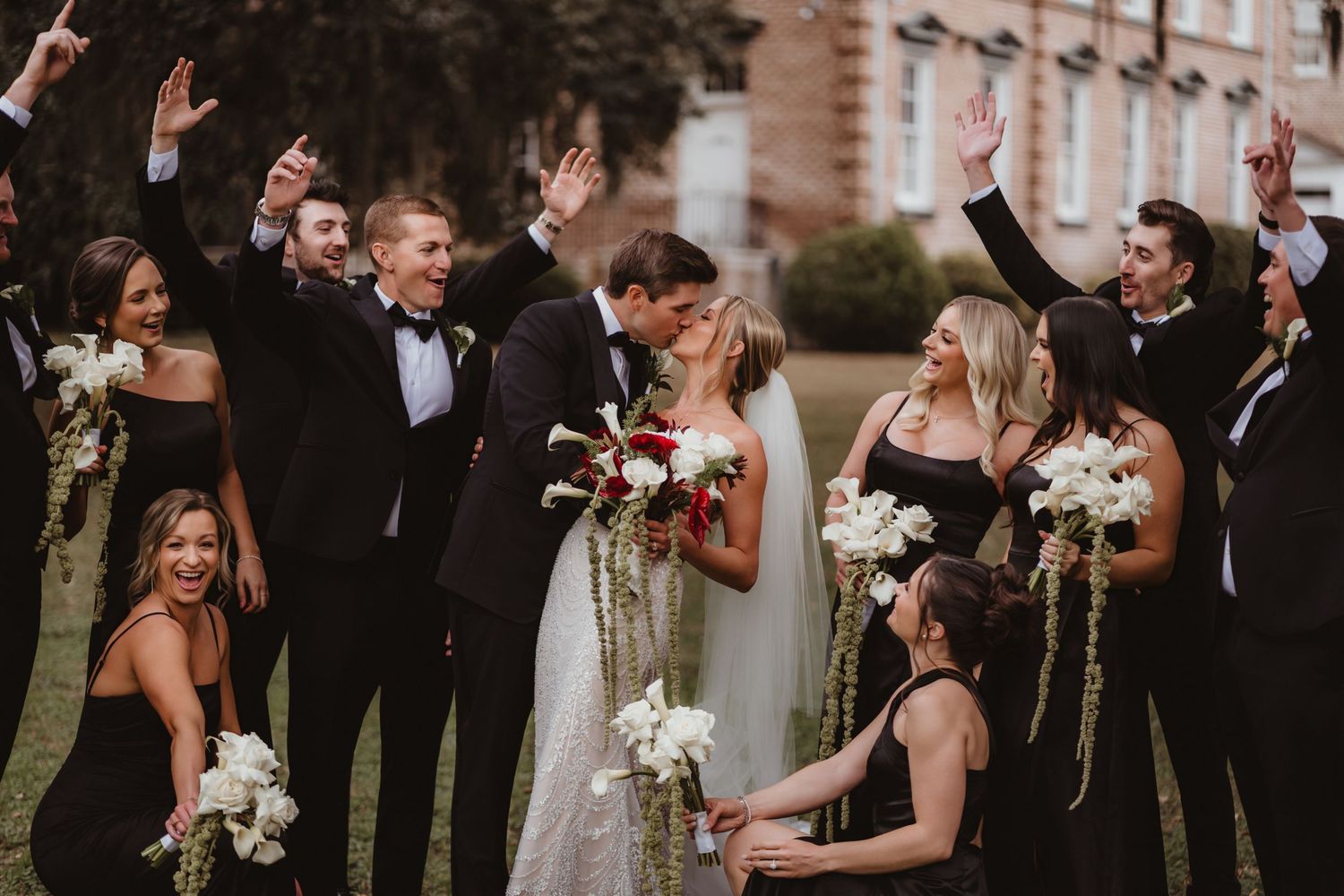 Bride and groom share a kiss surrounded by cheering wedding party in black attire holding white bouquets outdoors.