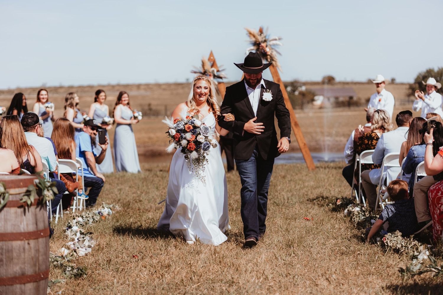 Bride and groom in black cowboy hat walk hand in hand down aisle at sunny outdoor country wedding.