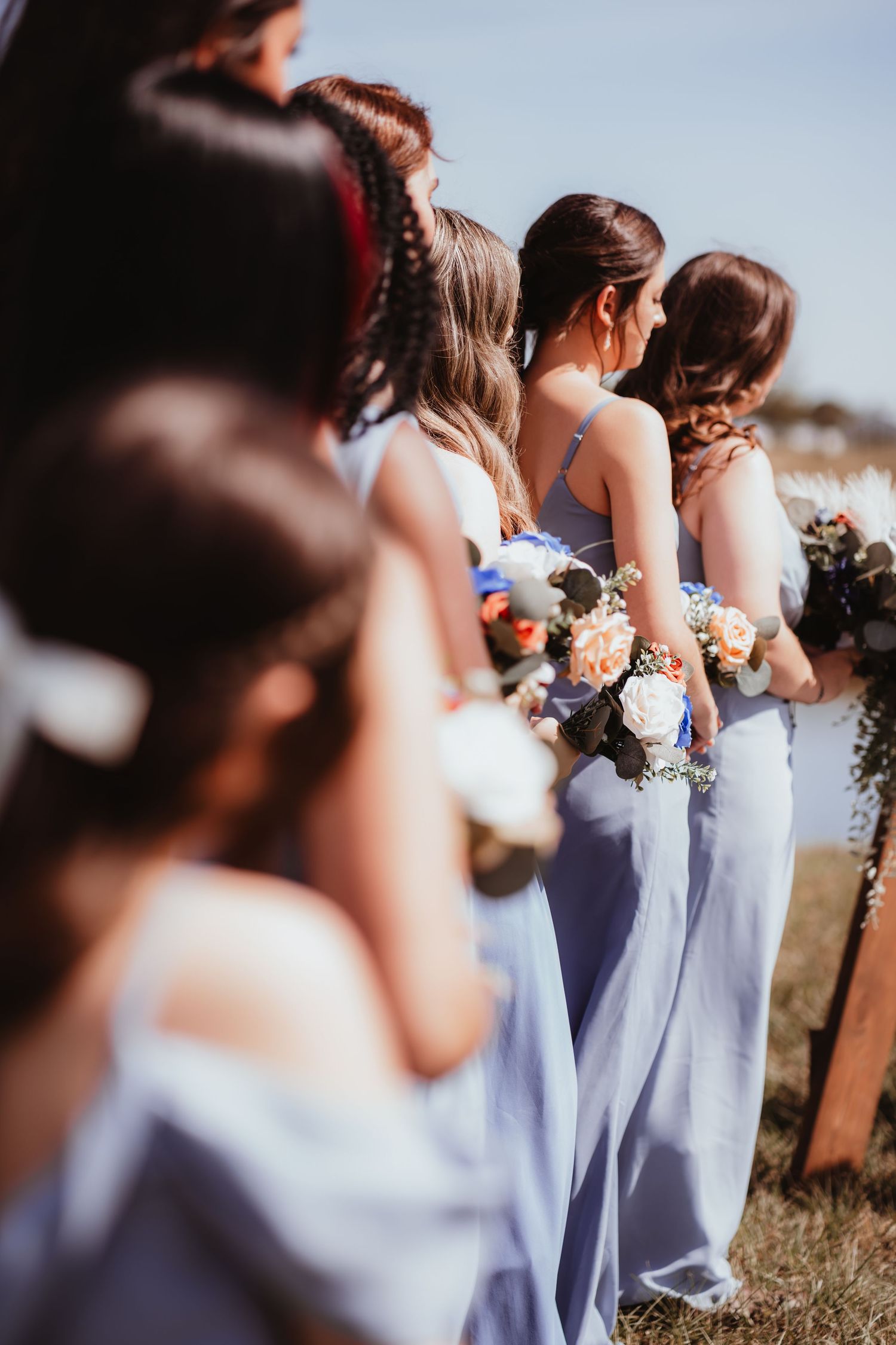 Bridesmaids in light blue dresses hold floral bouquets while standing in a row at an outdoor wedding ceremony.