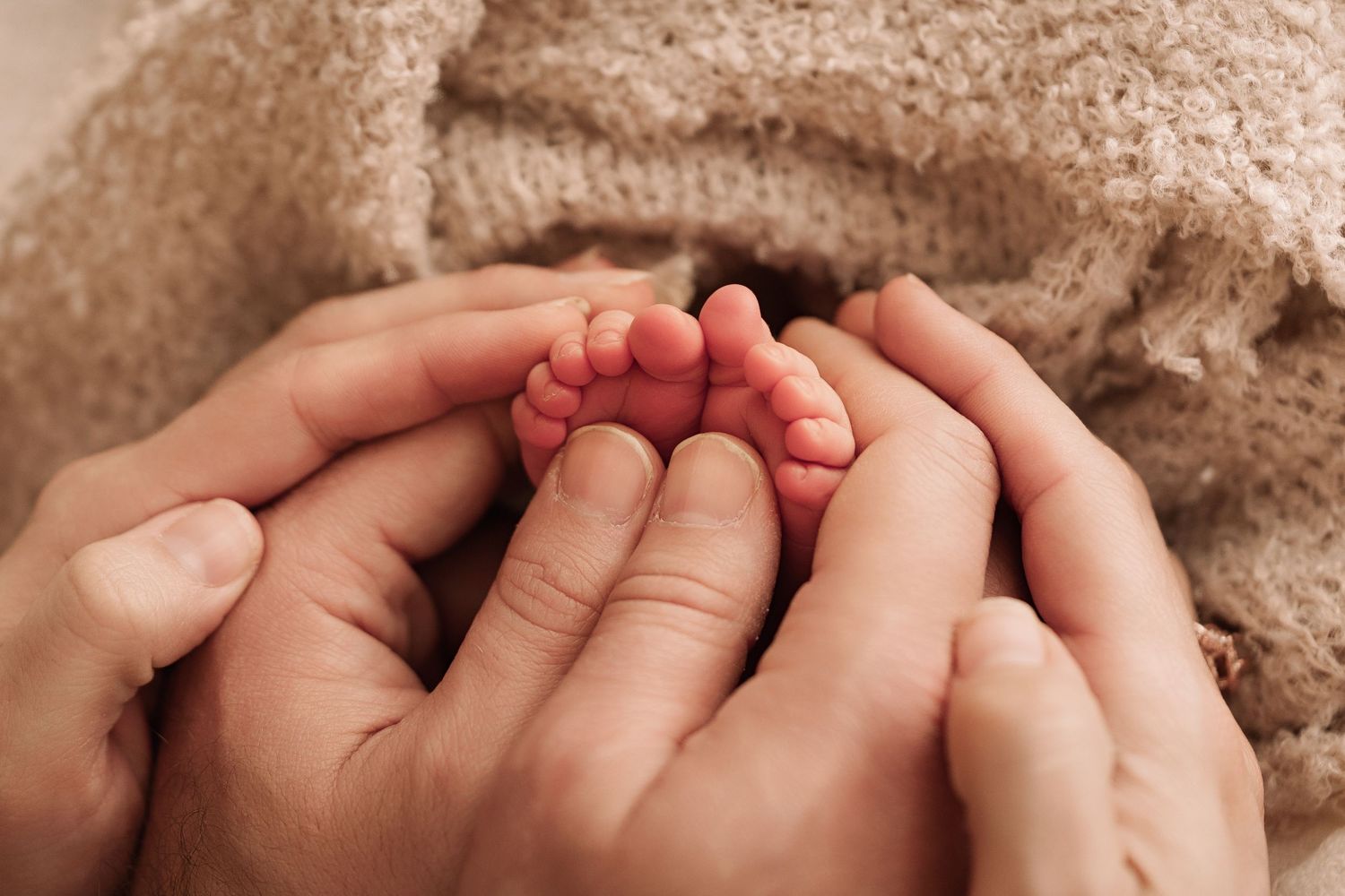 Two adult hands gently cradling tiny newborn baby feet wrapped in a soft beige blanket.