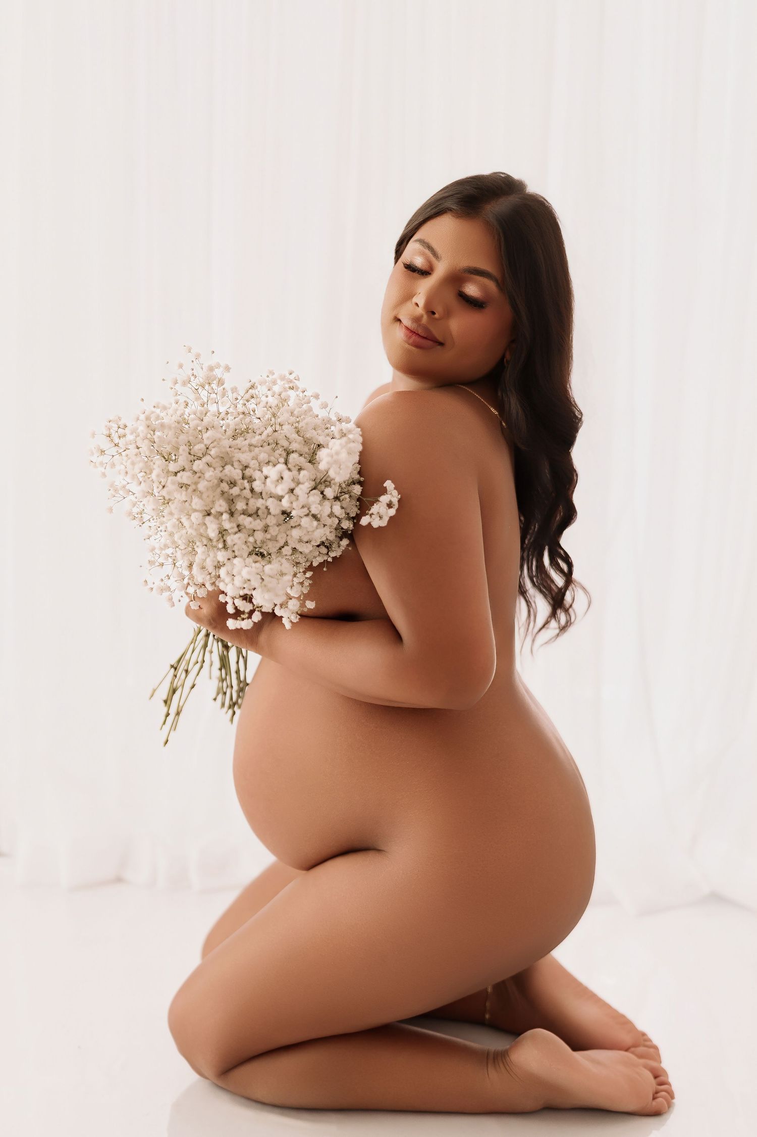 Nude pregnant woman kneeling while holding white flower bouquet in studio setting.