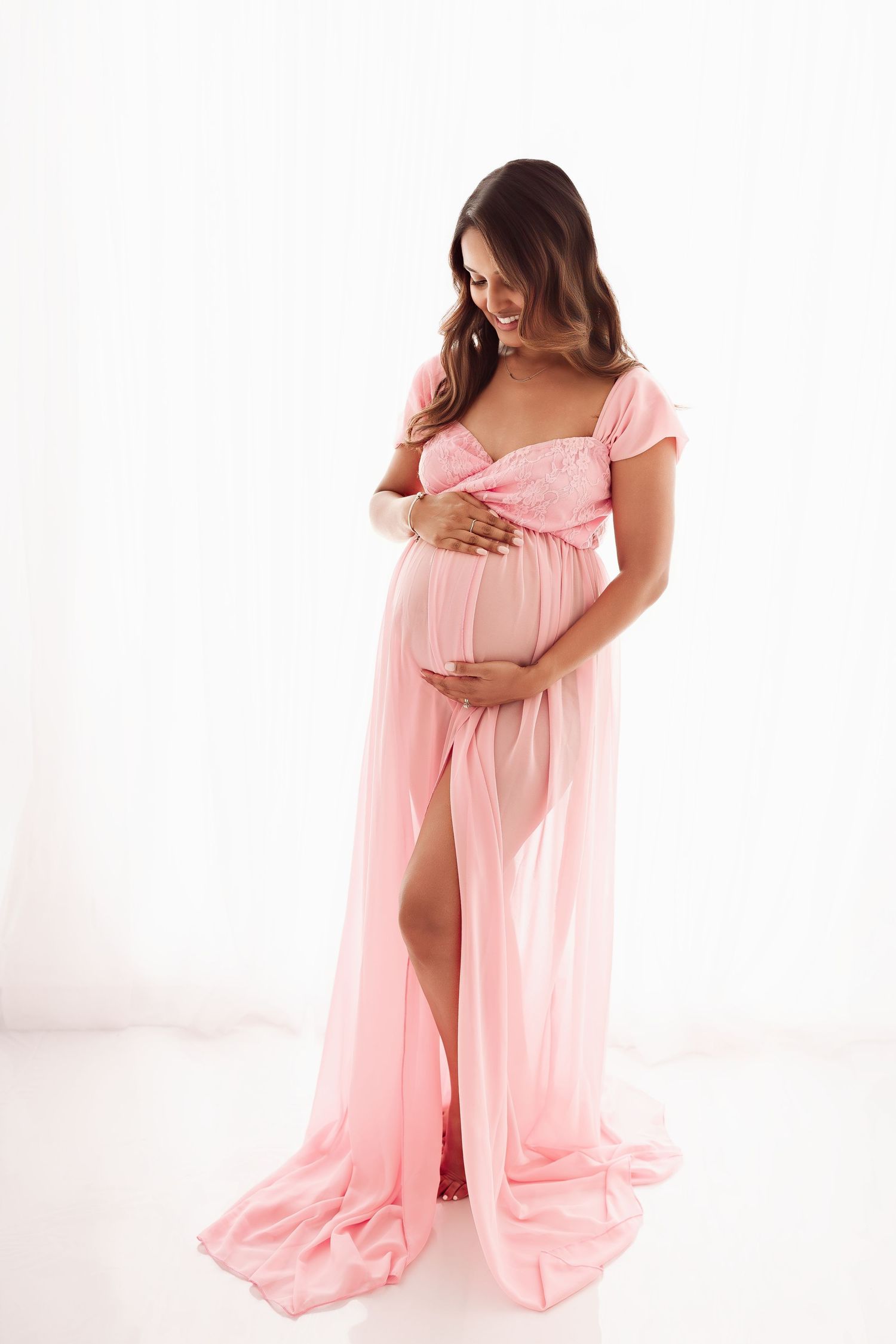 Pregnant woman in pink chiffon maternity gown cradling baby bump in white studio.