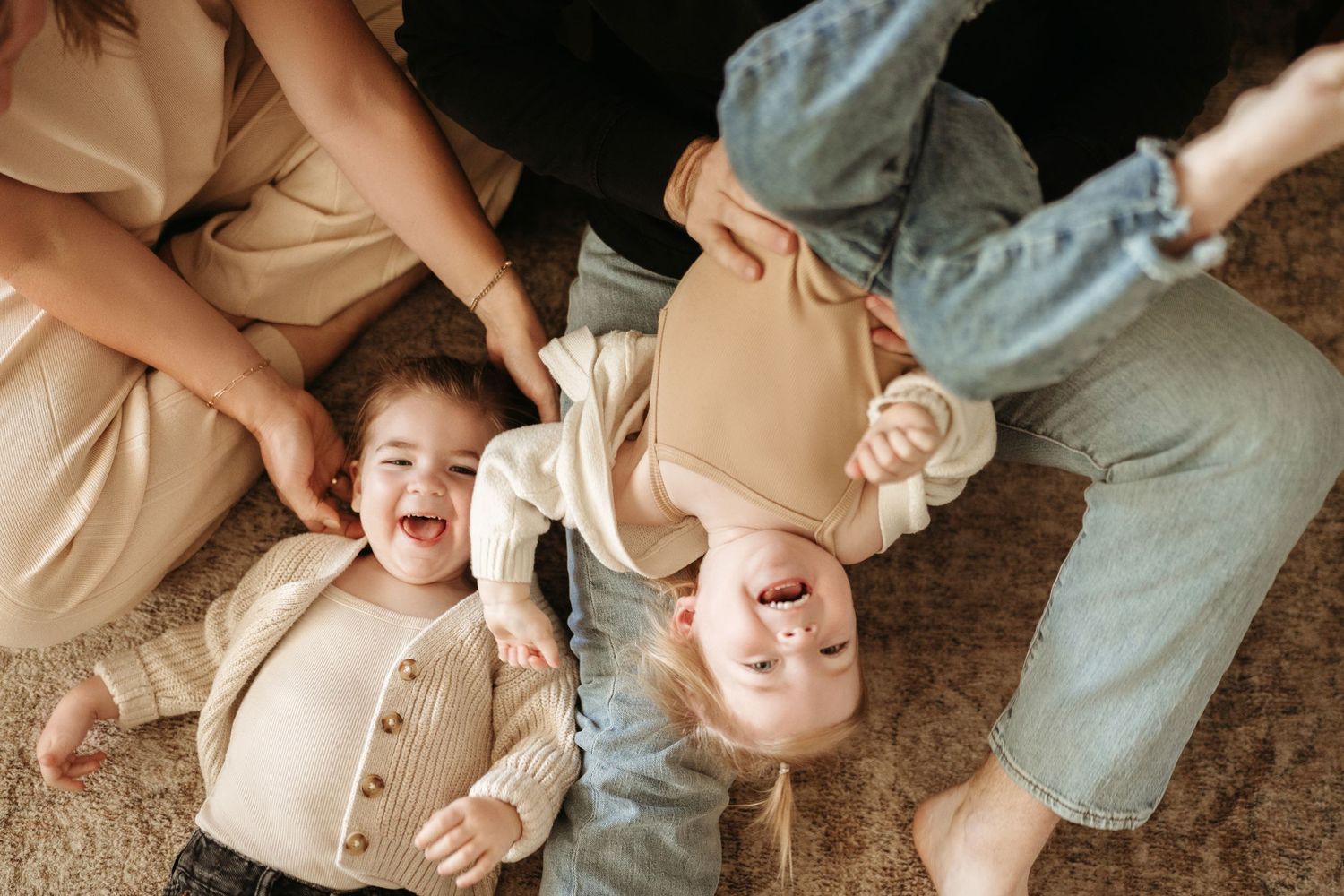 Overhead view of two young children laughing joyfully while lying on a carpet with parents nearby.