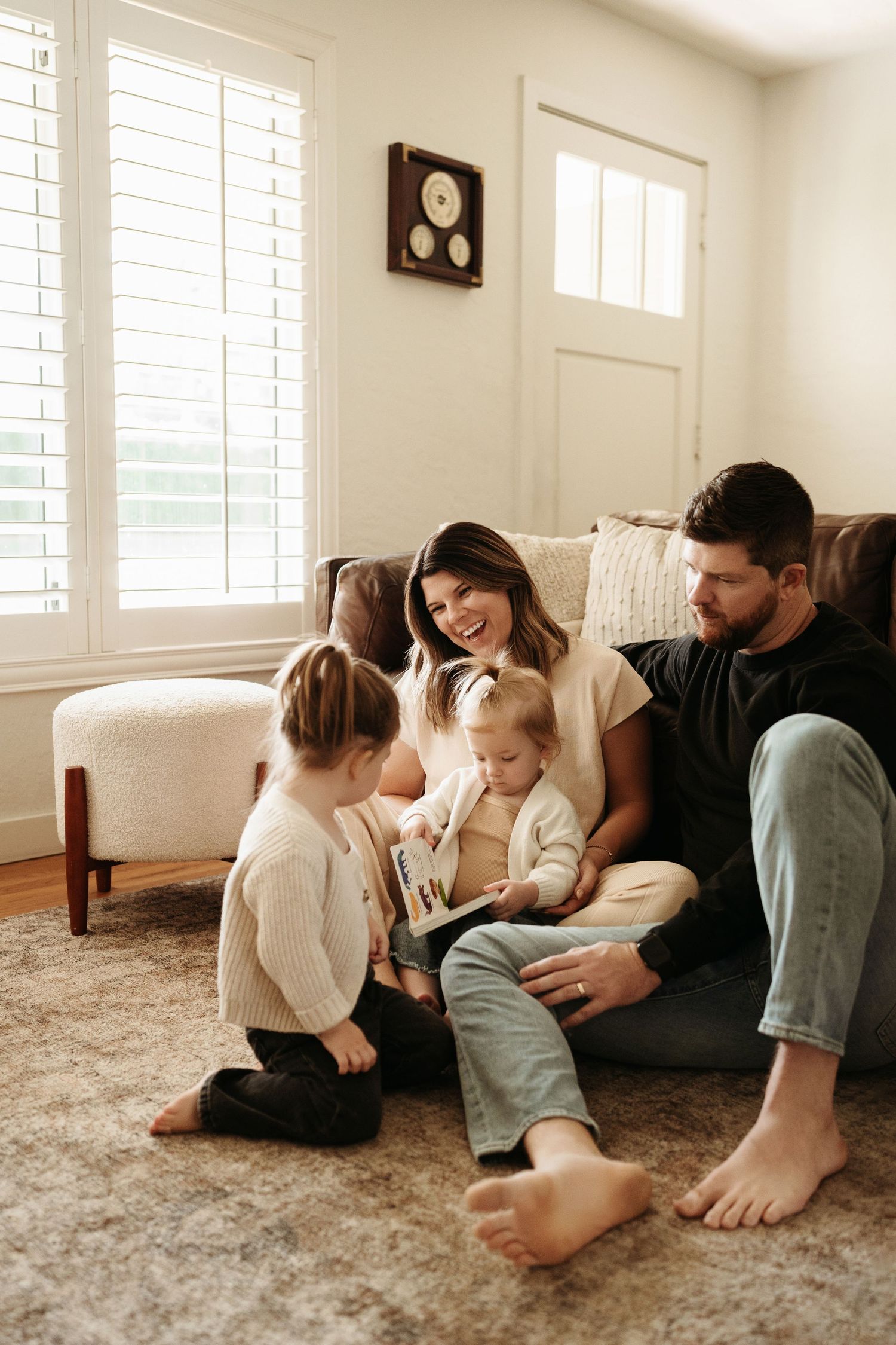 Family of four sharing a cozy moment reading together on a living room floor near a bright window.