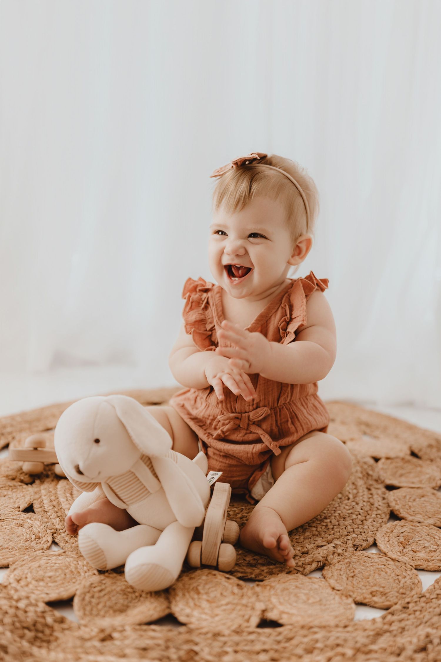 Laughing baby girl in terracotta romper clapping hands beside a white plush bunny on woven rug.