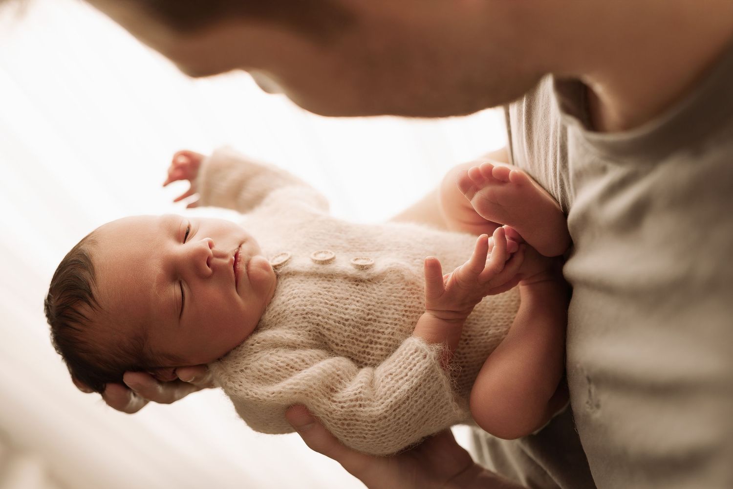 Father gently holding peaceful sleeping newborn baby in cream knit outfit near bright window.