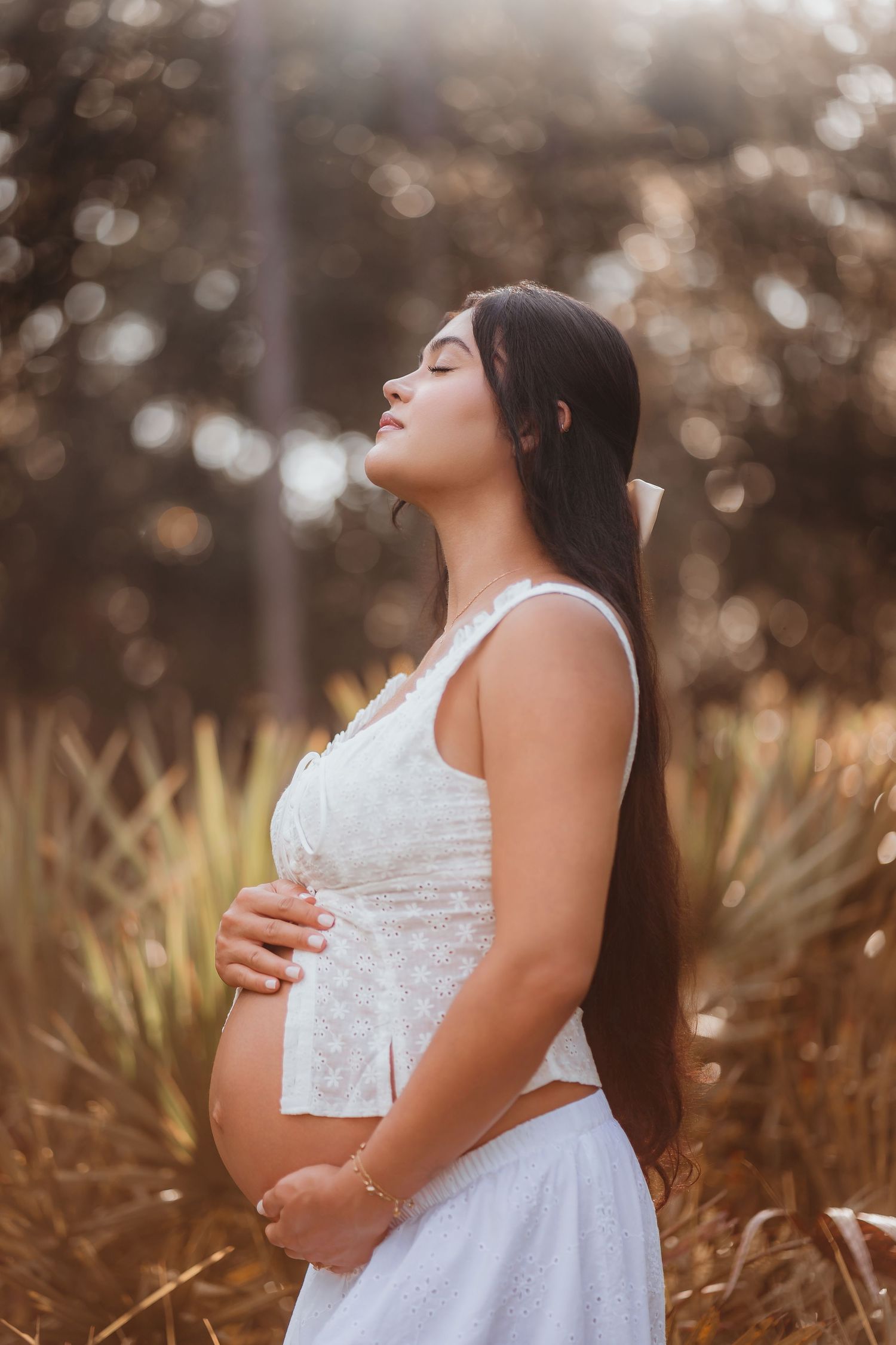 Serene pregnant woman in white dress holding baby bump with eyes closed in golden sunlit field.