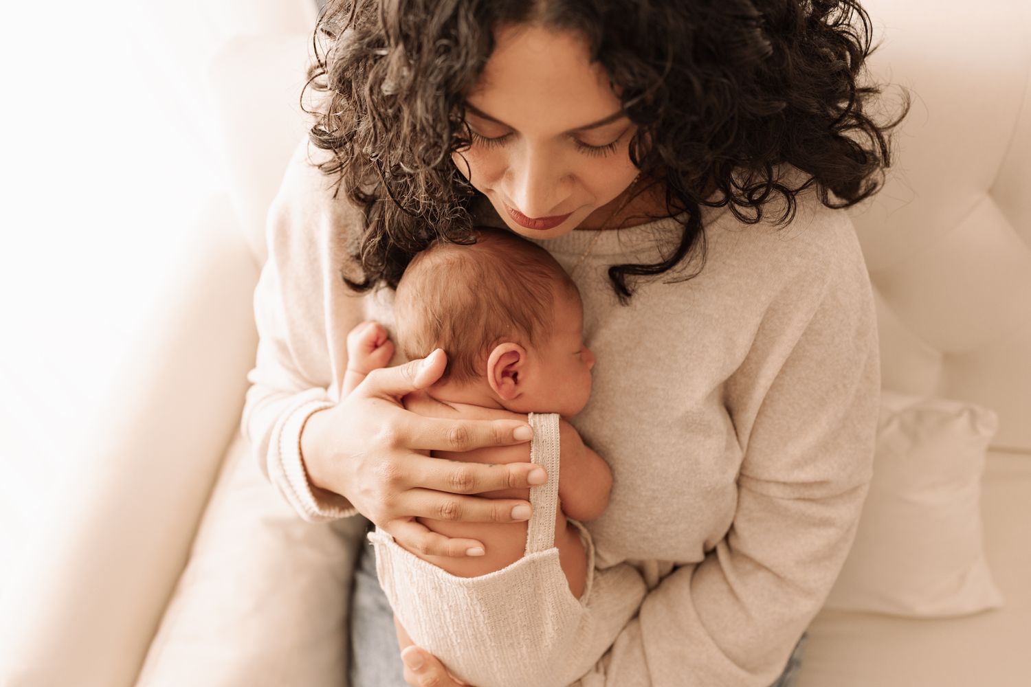 Mother with curly hair gently cradling and kissing her newborn baby indoors.