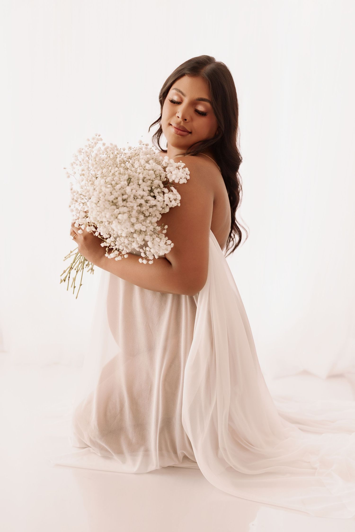 Pregnant woman in sheer white maternity dress holding bouquet of white flowers.