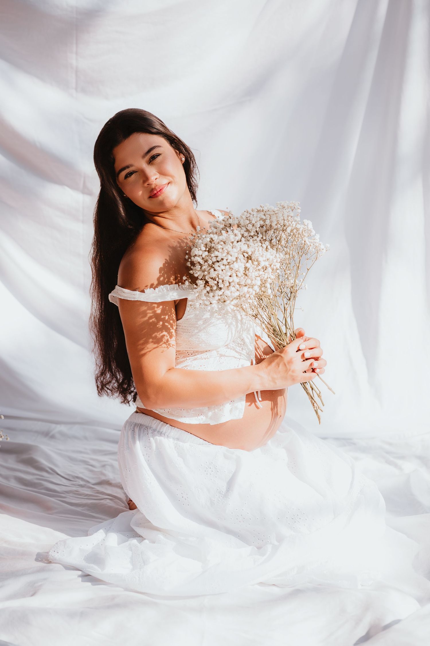 Pregnant woman in white dress clutching baby's breath flowers against flowing white fabric backdrop.