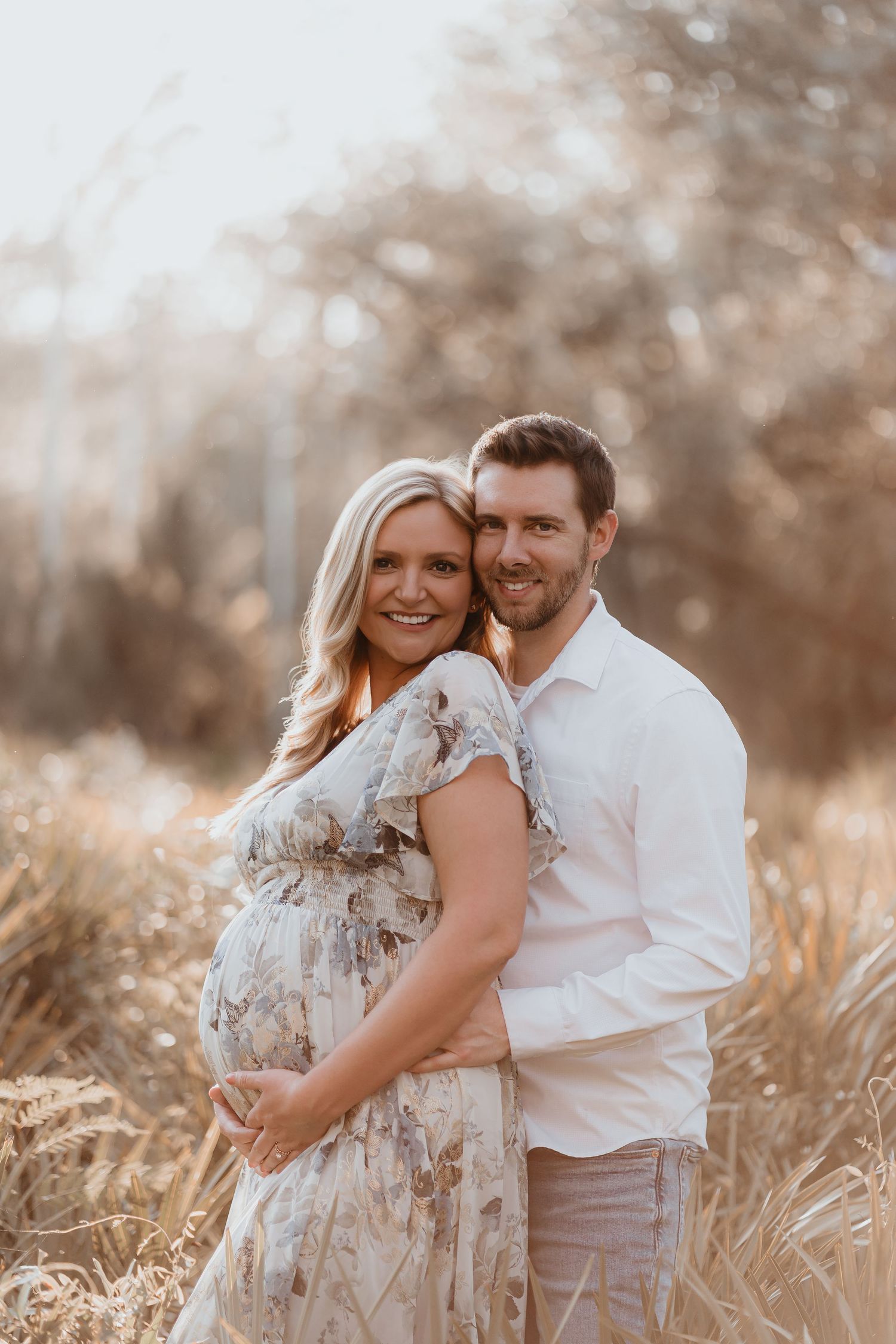 Smiling pregnant couple poses together in sun-drenched autumn field, woman in floral maternity dress.