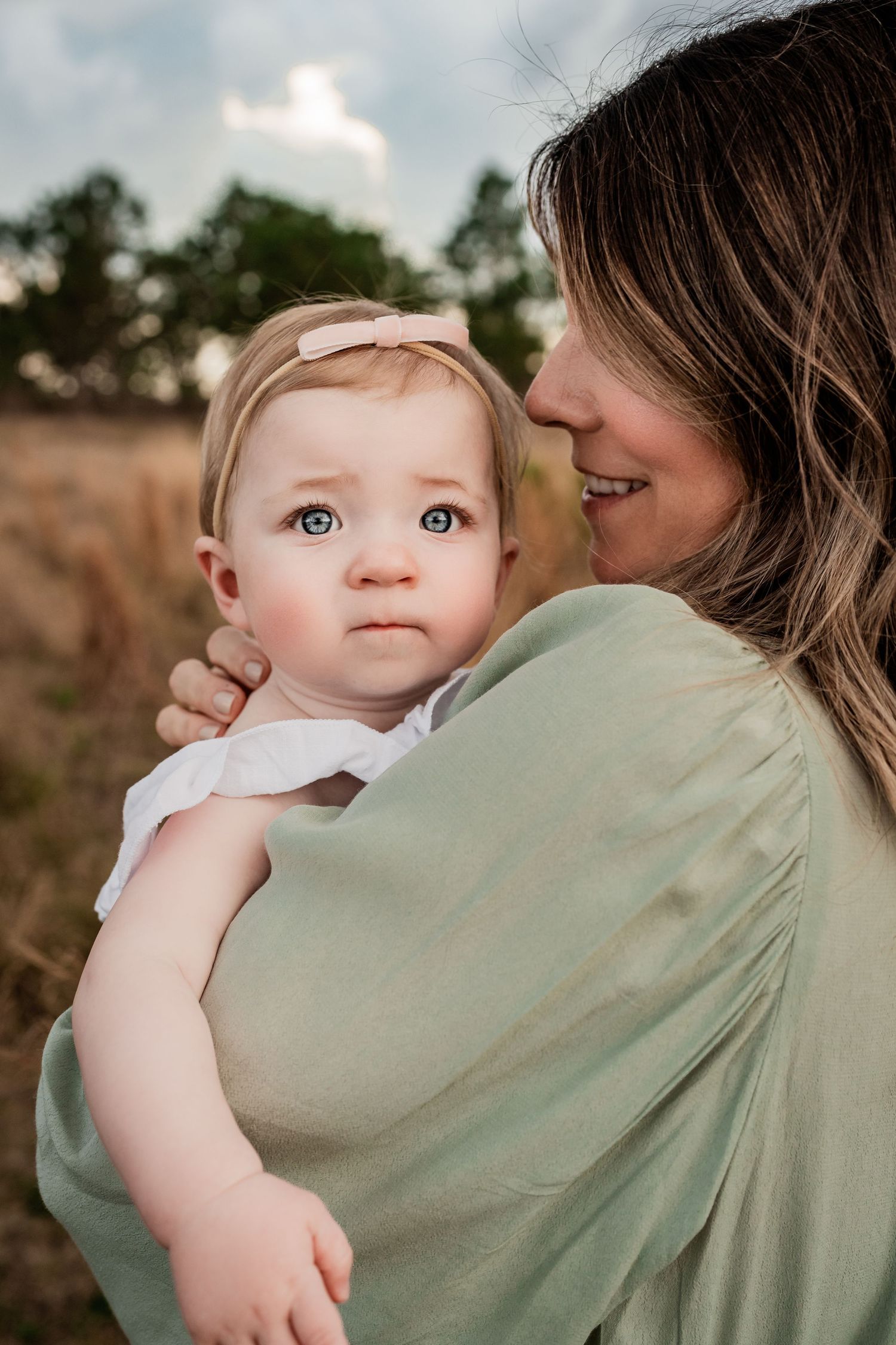 Sweet baby girl with pink bow headband gazing forward while mother holds her in a field.