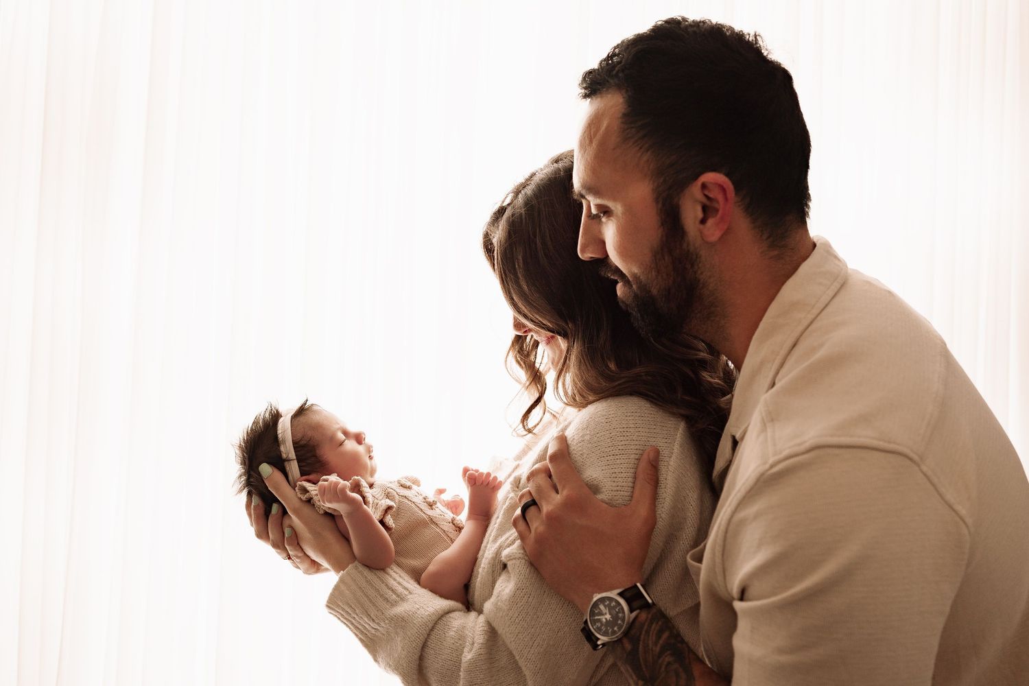 A couple shares a tender moment together while cradling their newborn baby near a bright window.