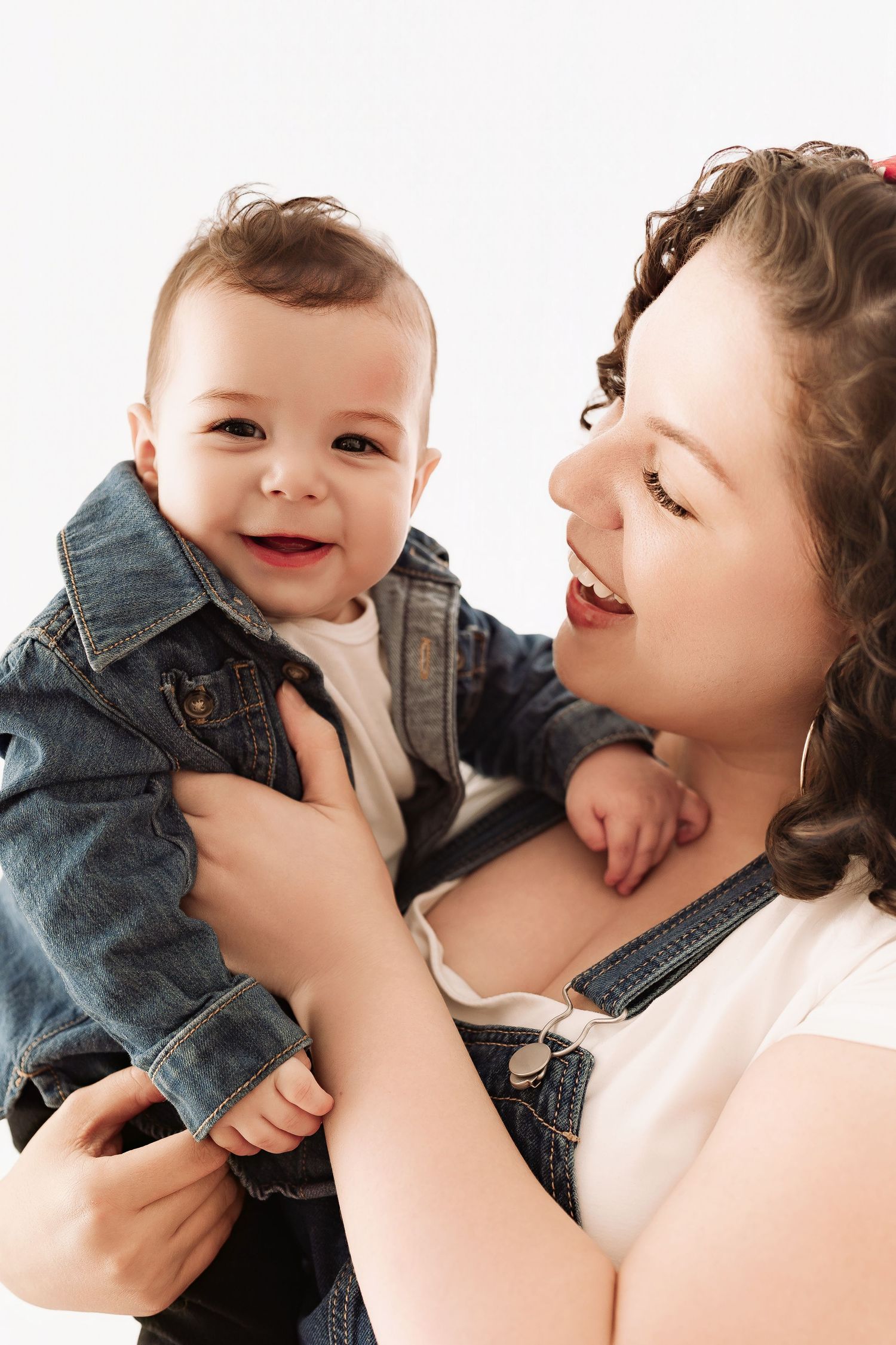 Joyful baby in denim jacket smiling brightly while being held by his curly-haired mother.