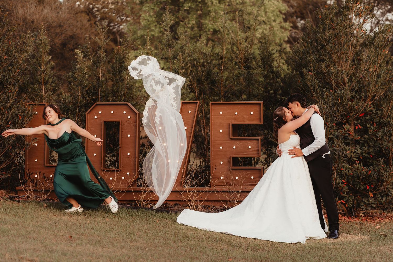 Excited bridesmaid throwing bridal veil into air beside kissing couple at LOVE sign backdrop.