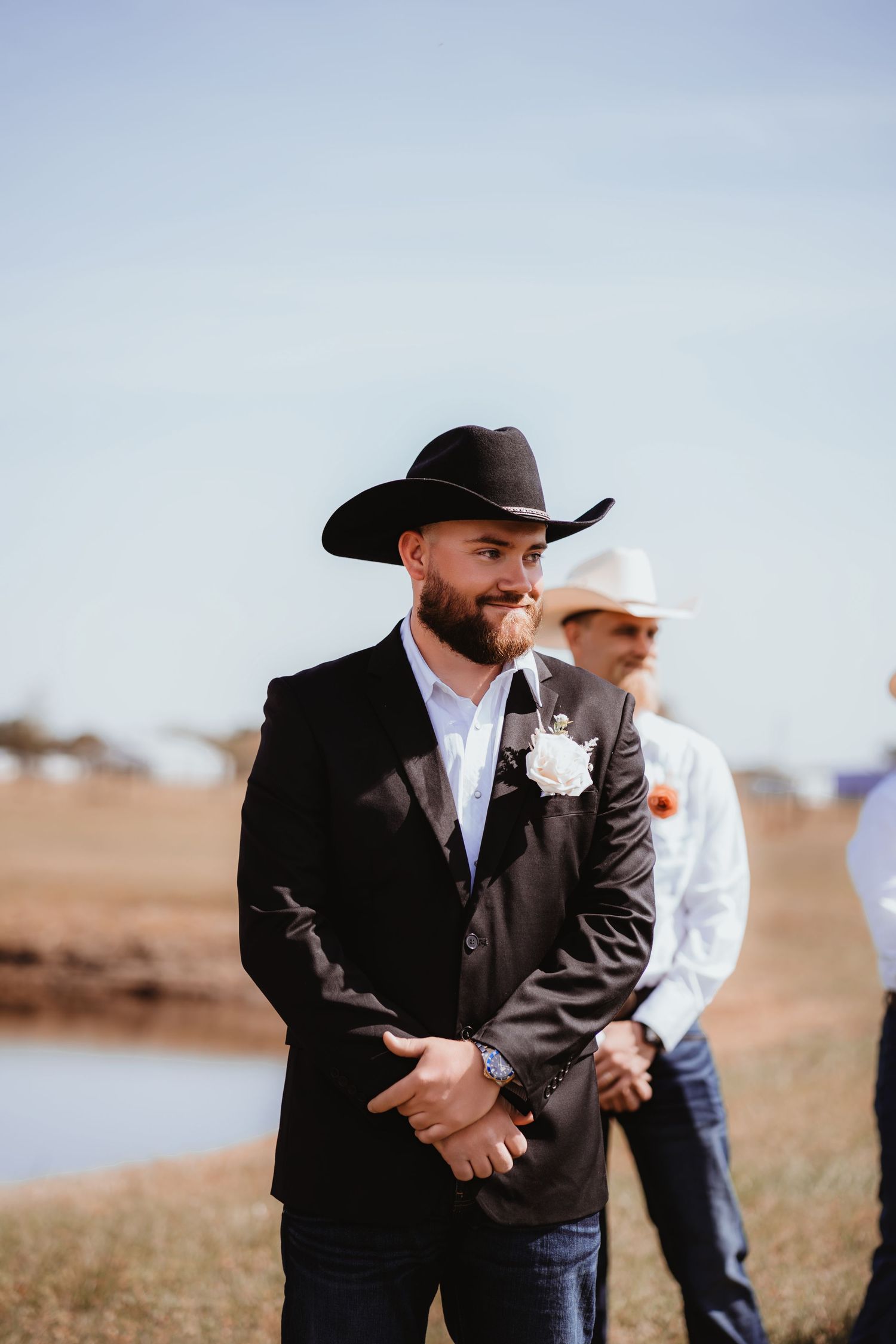 Handsome groom in black cowboy hat and dark blazer smiles as bride walks down the aisle outdoors.