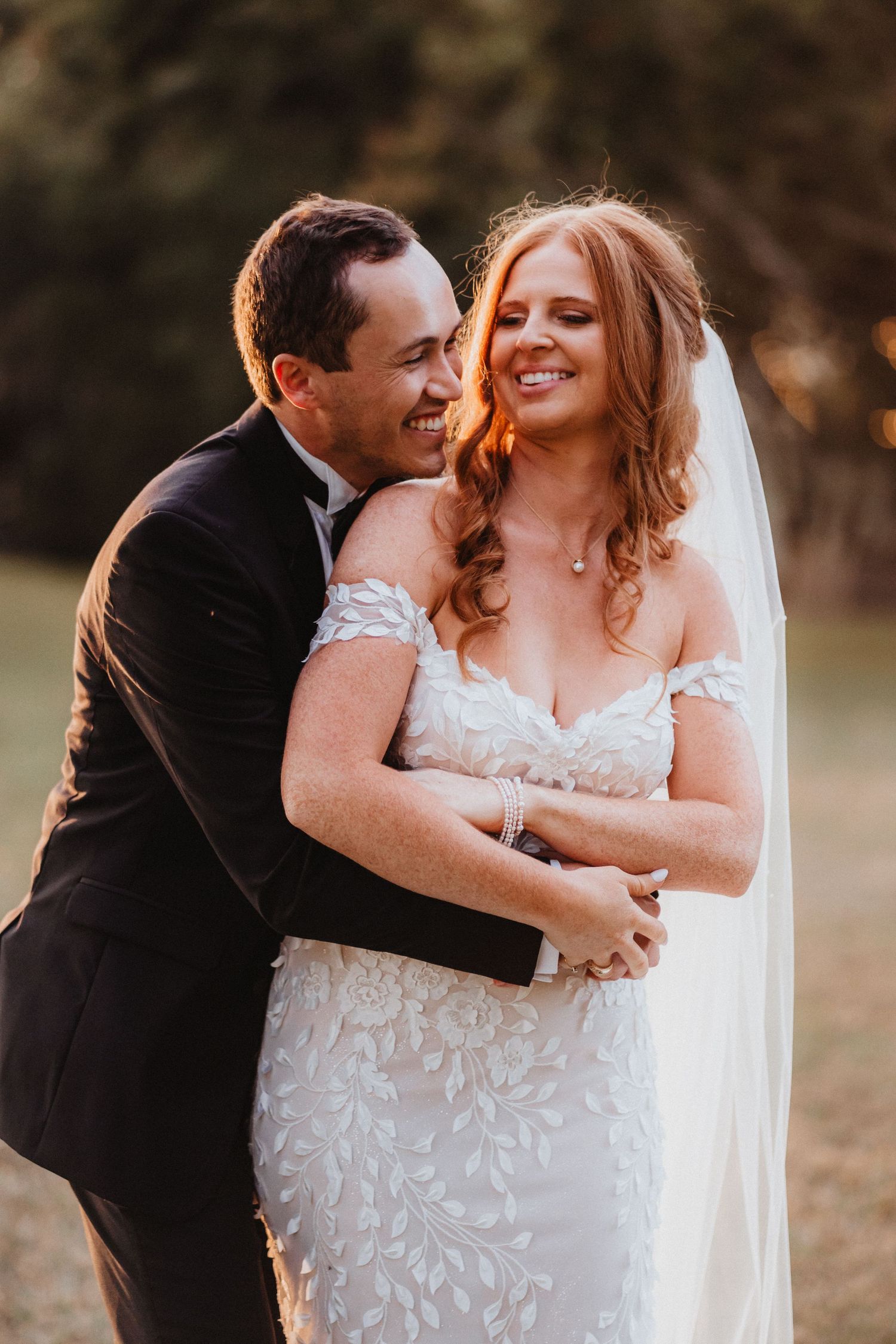 Groom embraces laughing bride from behind outdoors, bride wearing lace off-shoulder mermaid gown and long veil.