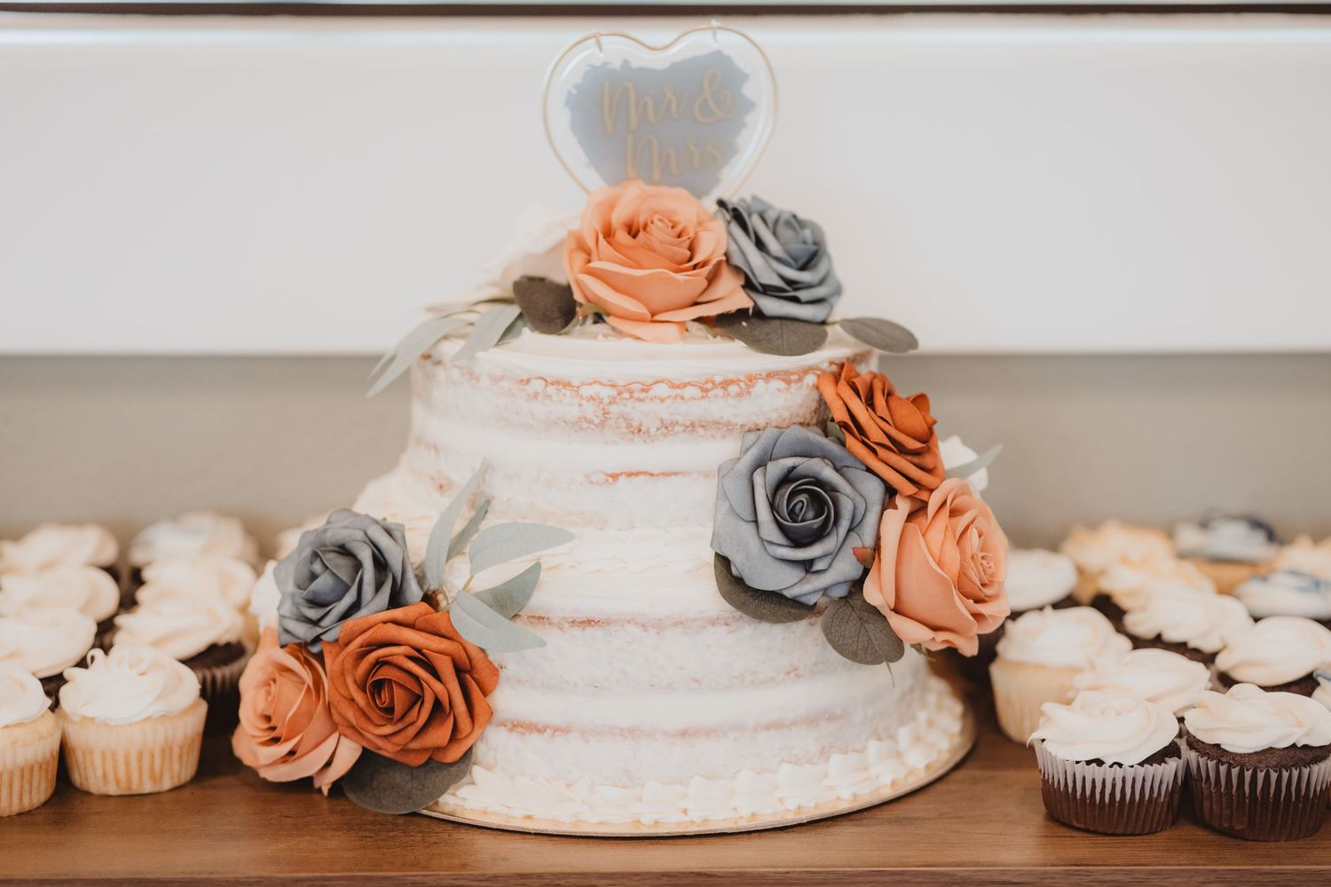 Two-tier naked wedding cake with orange and gray roses, heart topper, surrounded by assorted cupcakes on wooden table.
