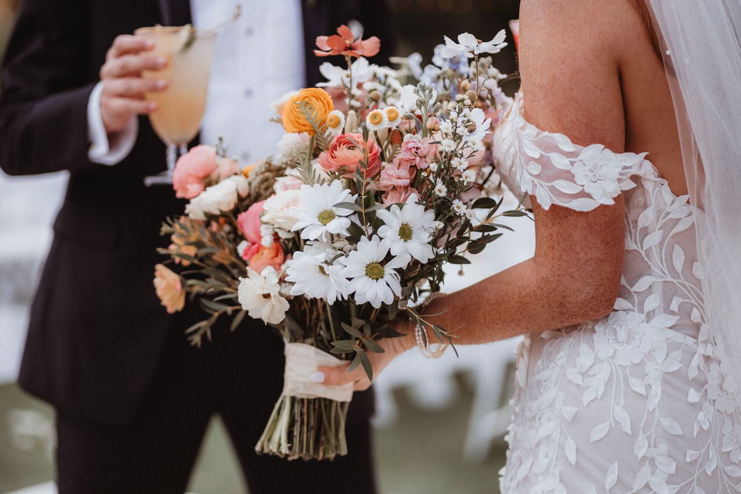 Bride holds a colorful wildflower bouquet with daisies, roses, and greenery on her wedding day.