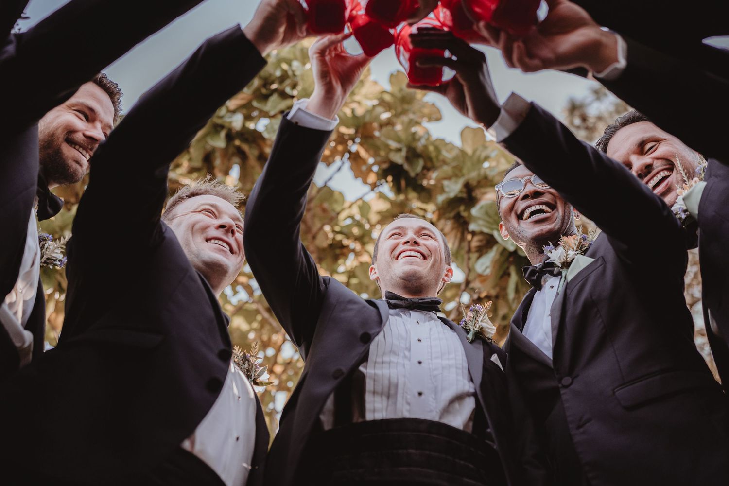 Groomsmen in black tuxedos raising red cups together in a low-angle celebratory wedding toast shot.