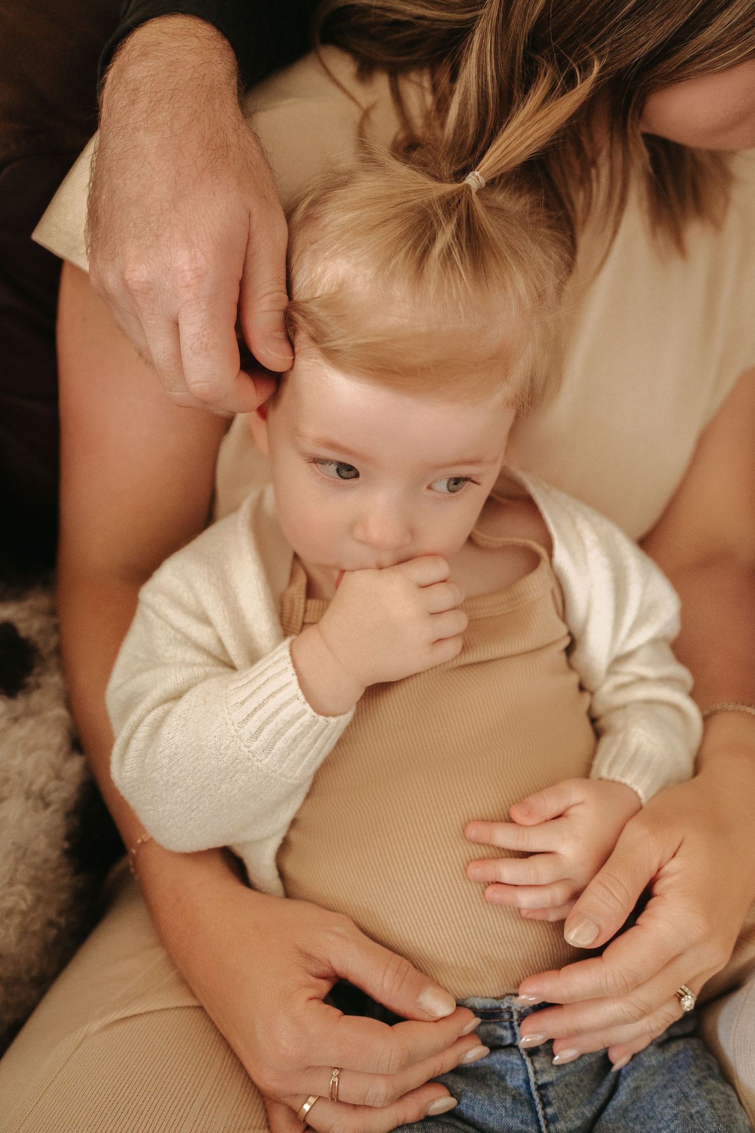 A blonde toddler in a cream cardigan sits contentedly in a mothers lap sucking fingers.