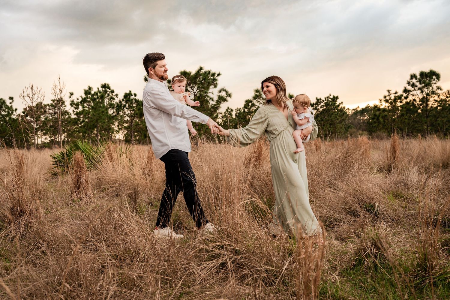 Young family walking through a golden field at sunset, parents each holding a baby girl in warm light.