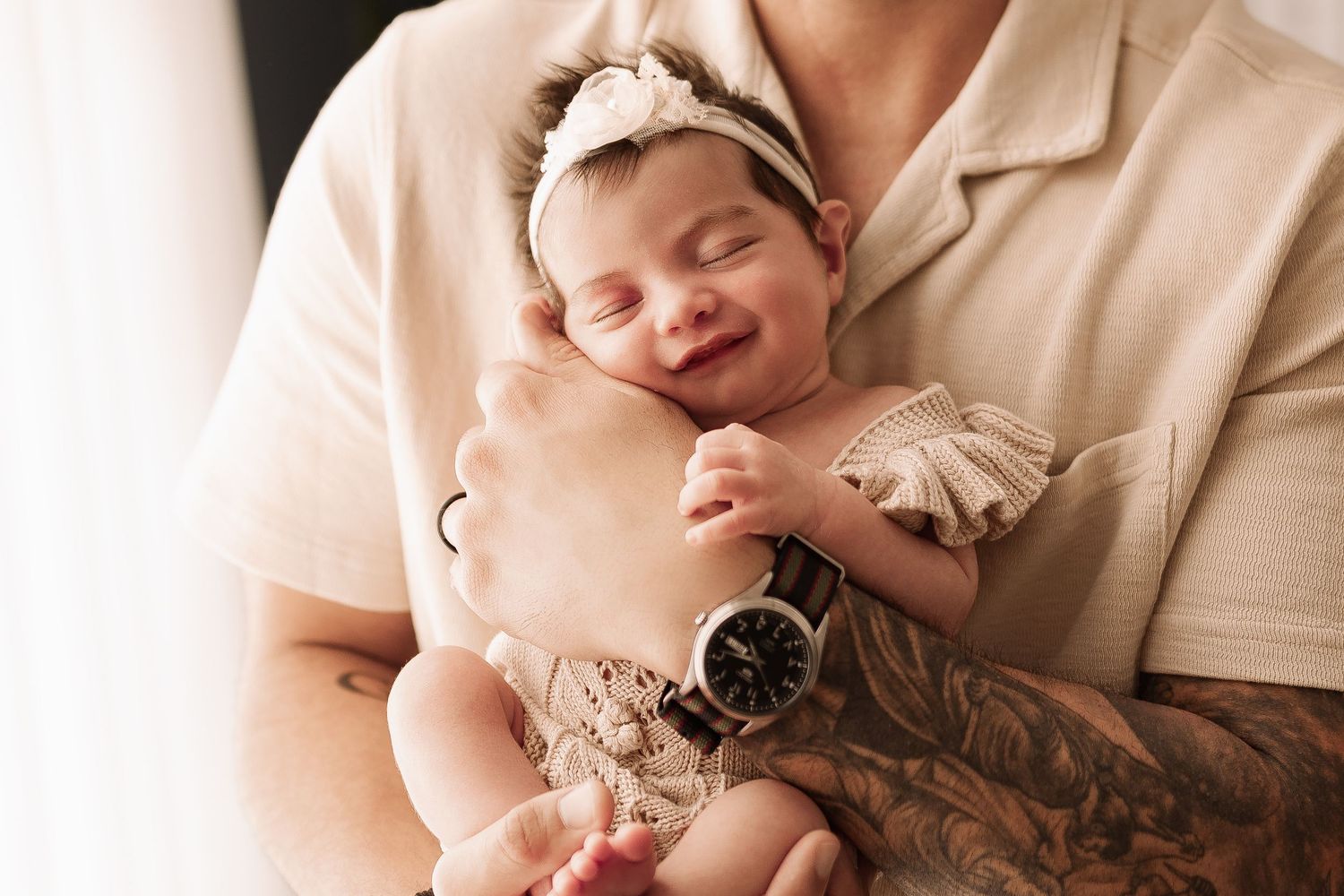 Close-up of sleeping newborn baby girl smiling in fathers tattooed arms wearing floral headband.
