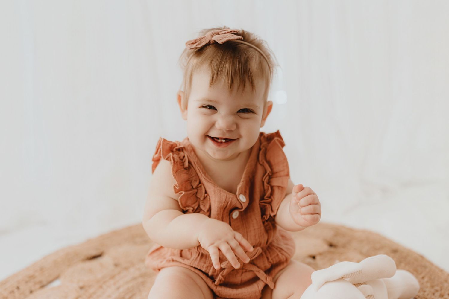 Smiling baby girl in rust orange ruffle romper with bow headband sitting on woven rug.