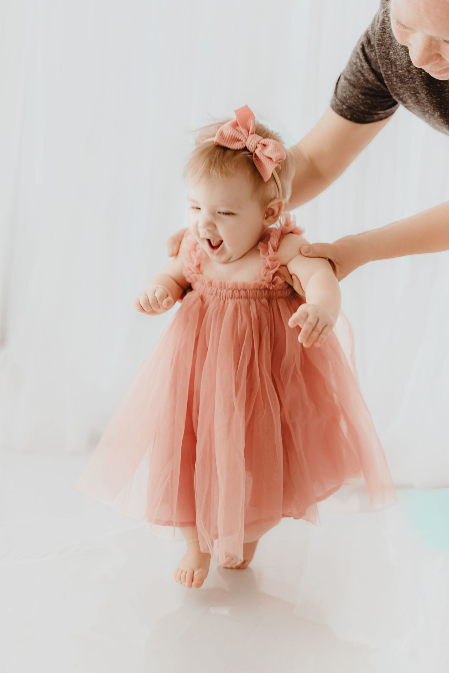 Baby girl in pink tulle dress and bow headband practices standing.