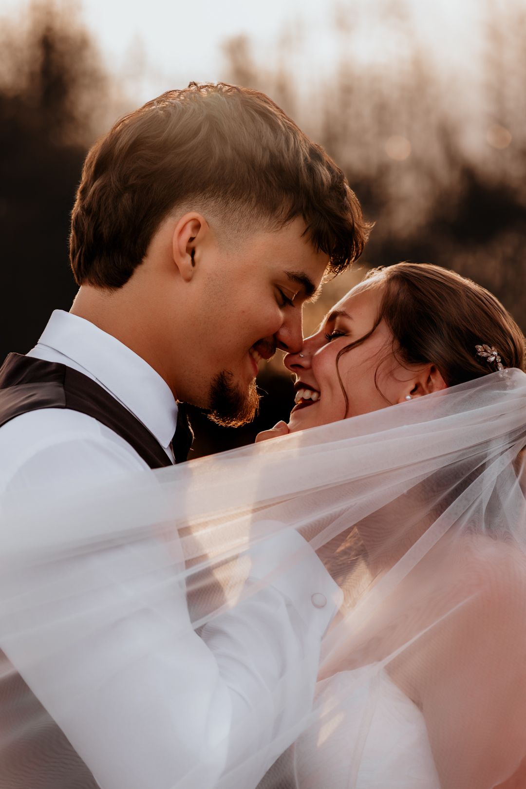 Laughing bride and groom sharing a joyful moment under a flowing veil in warm sunset light.