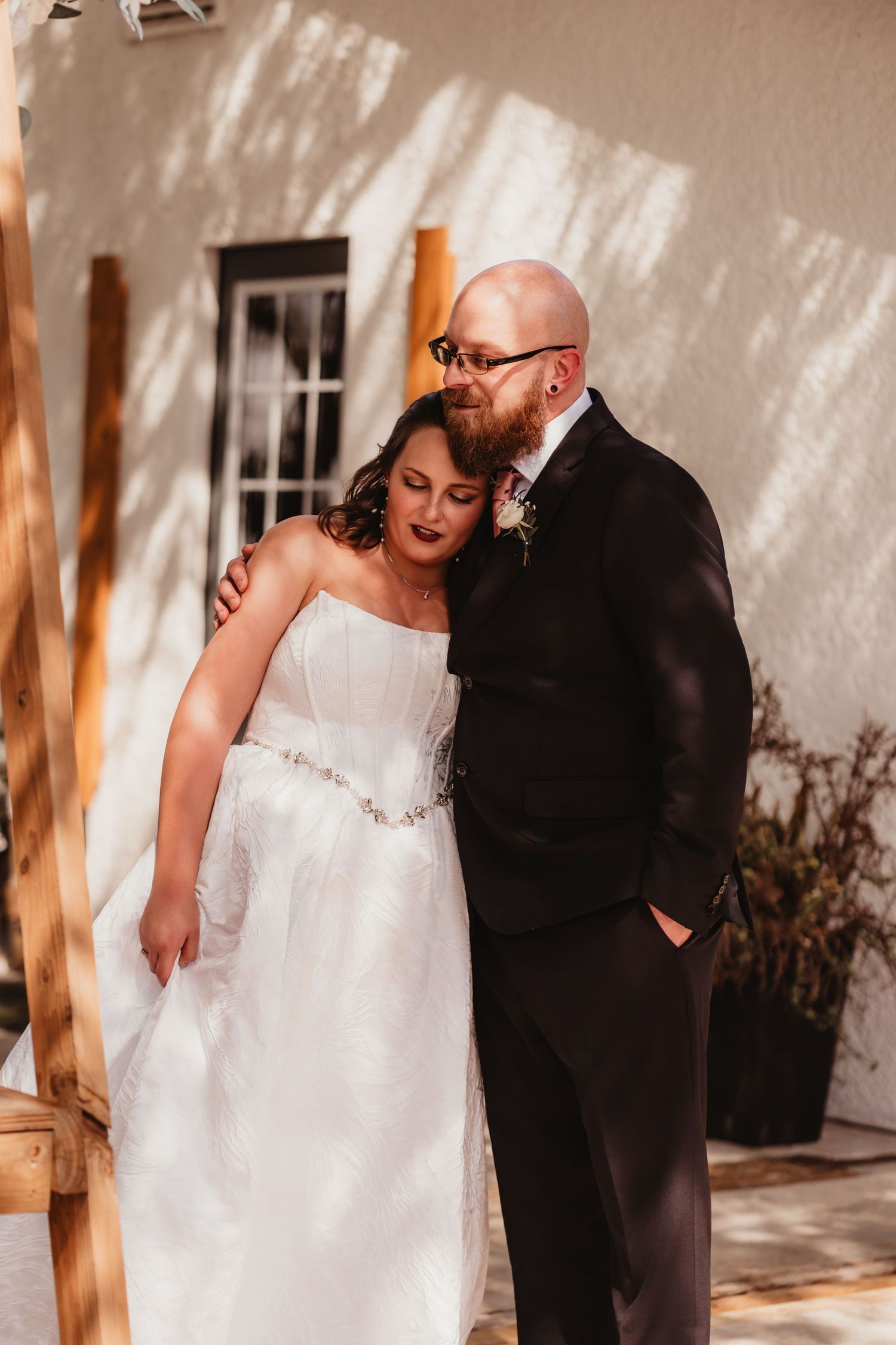 Bald groom with beard in black suit tenderly embracing bride in strapless white gown against white wall.