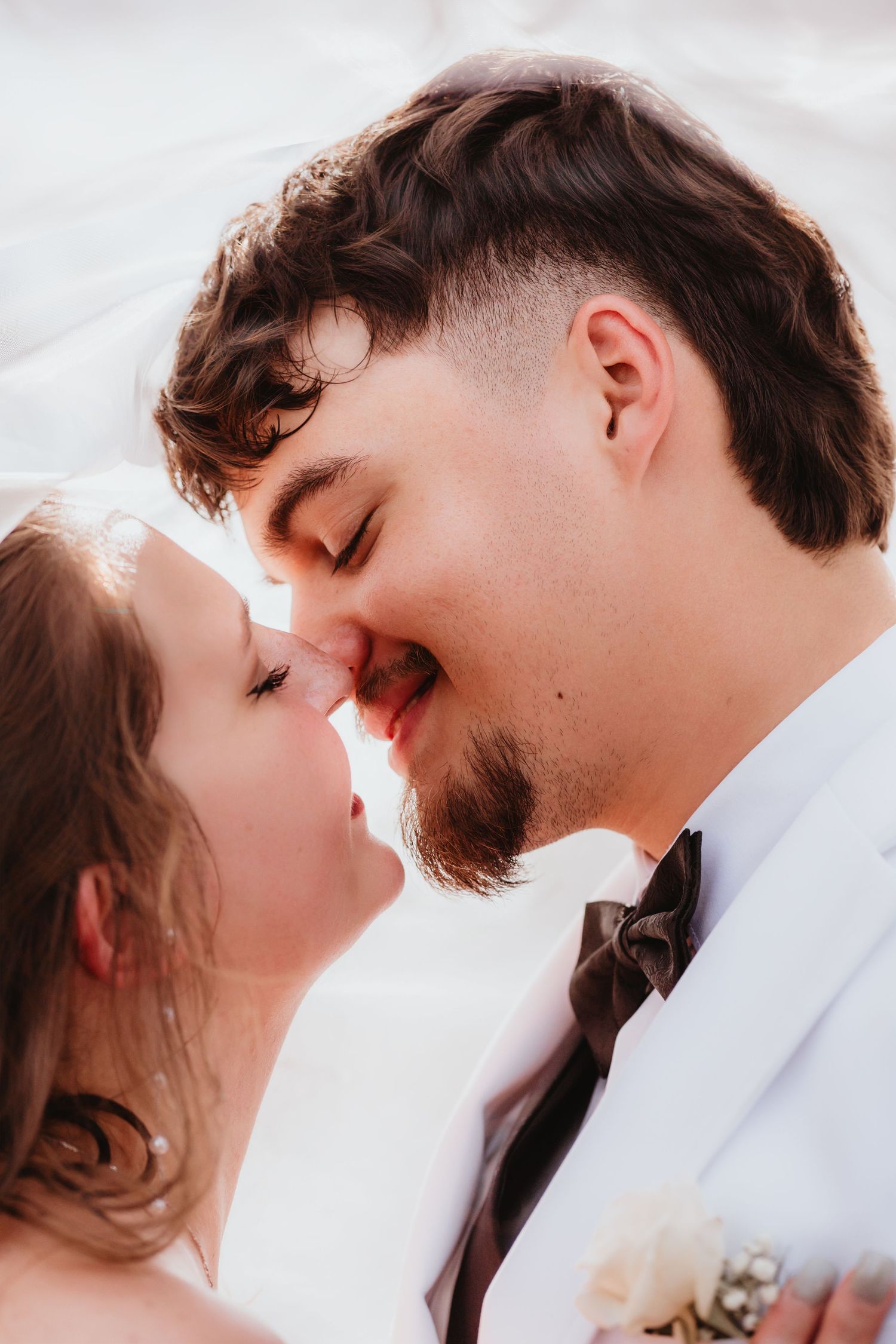 Smiling groom in white tuxedo leaning in to kiss his bride beneath a sheer wedding veil.