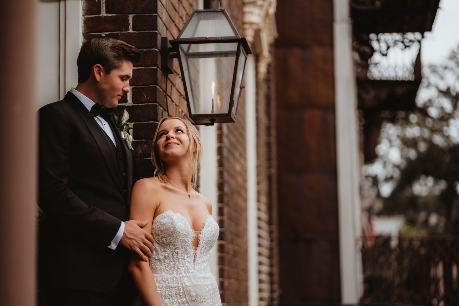 Bride in a plunging lace gown gazes up at groom in black tuxedo beside a brick wall with vintage lantern.