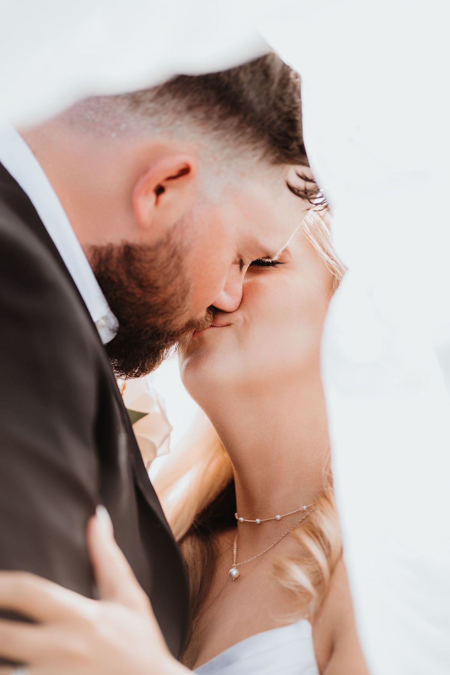 Soft romantic close-up of newlyweds kissing under a veil, bride wearing a delicate pearl necklace and earrings.