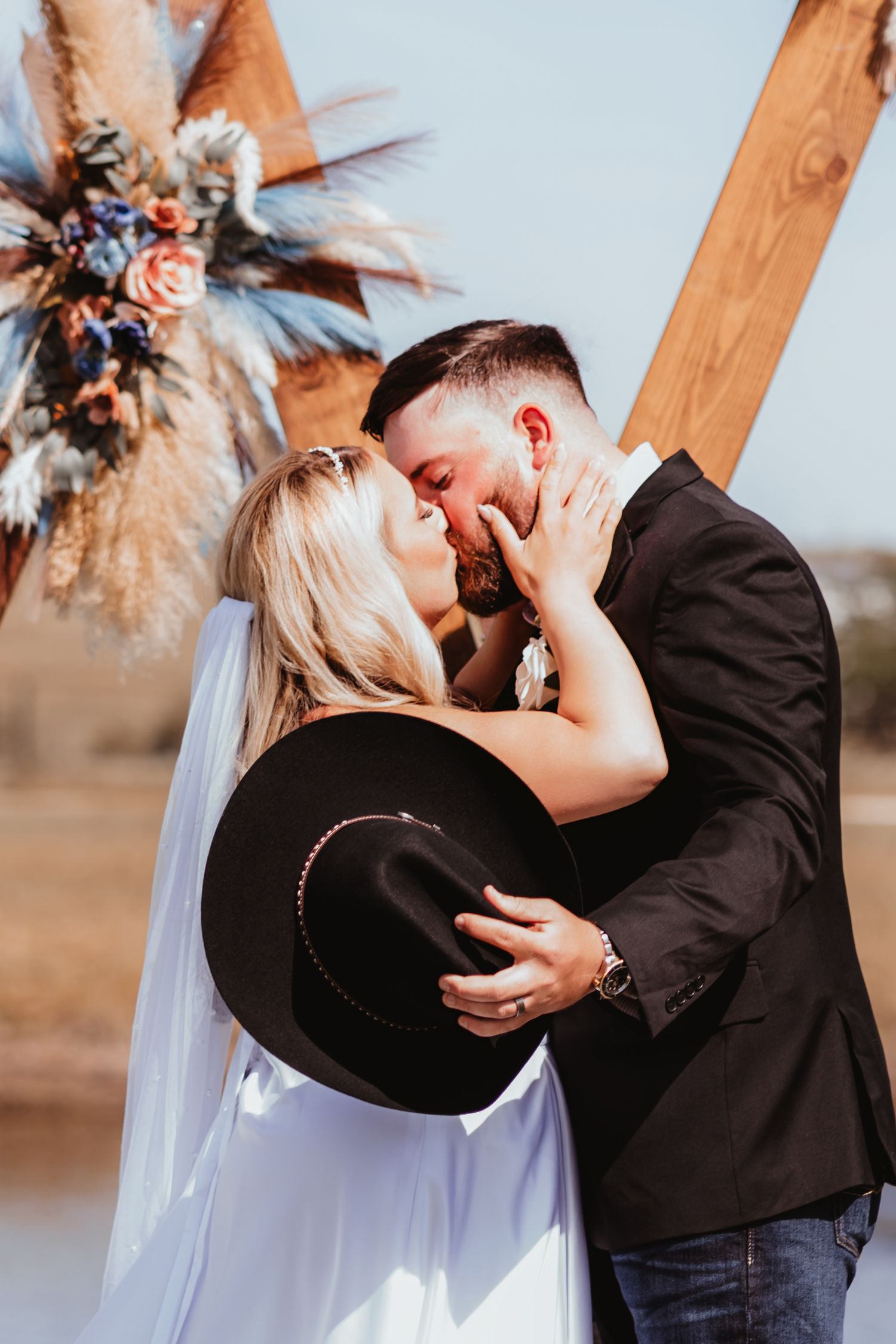 Bride and groom share first kiss at boho outdoor wedding, bride holding groom's black cowboy hat.
