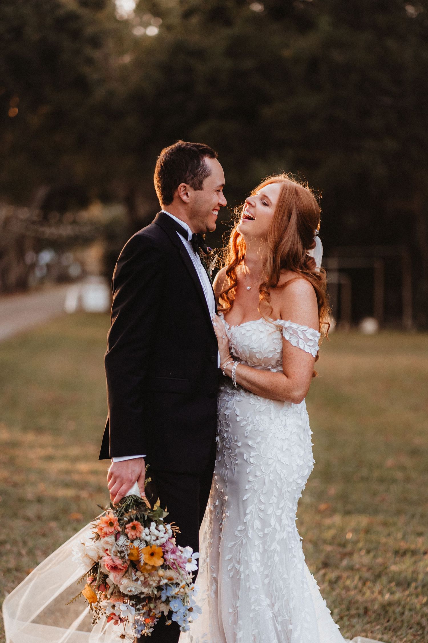 Laughing bride in lace off-shoulder gown and groom in tuxedo share a joyful moment during golden hour.